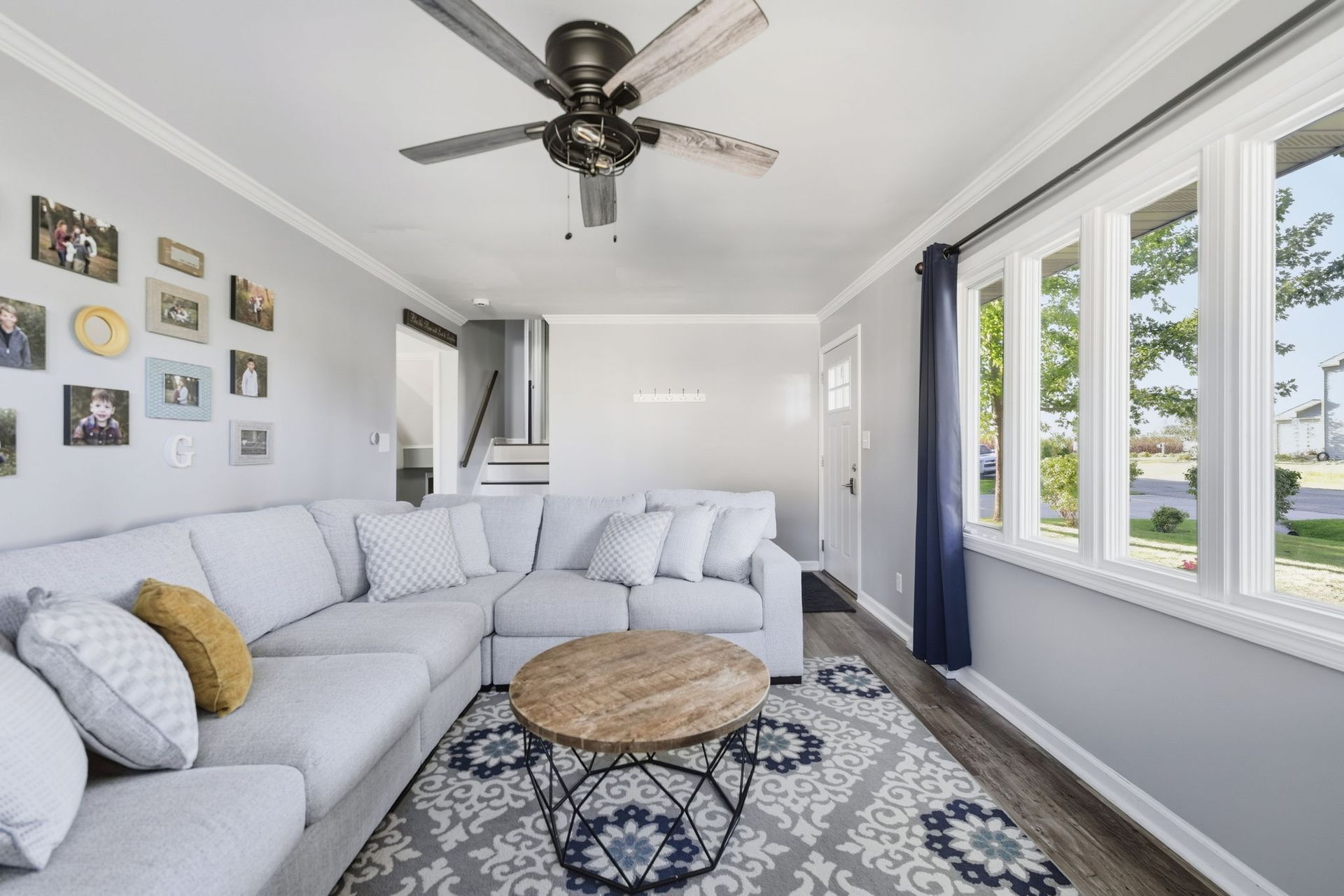 Bright living room with a gray sectional, patterned rug, ceiling fan, and large windows.