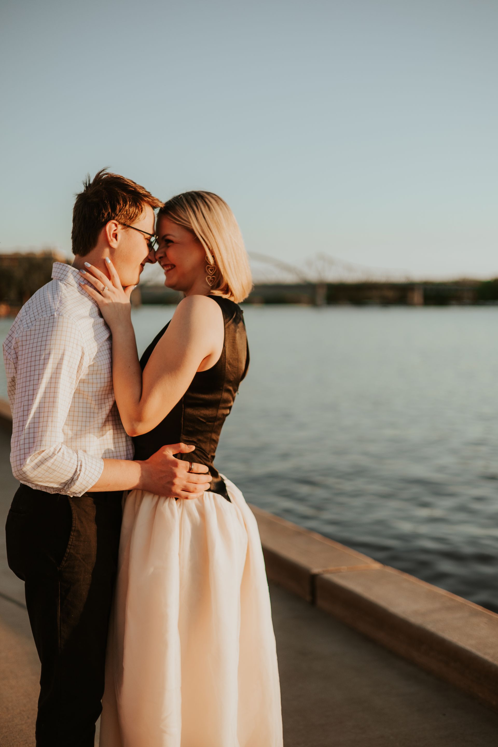 Couple embracing by the water at sunset, with a bridge in the background.