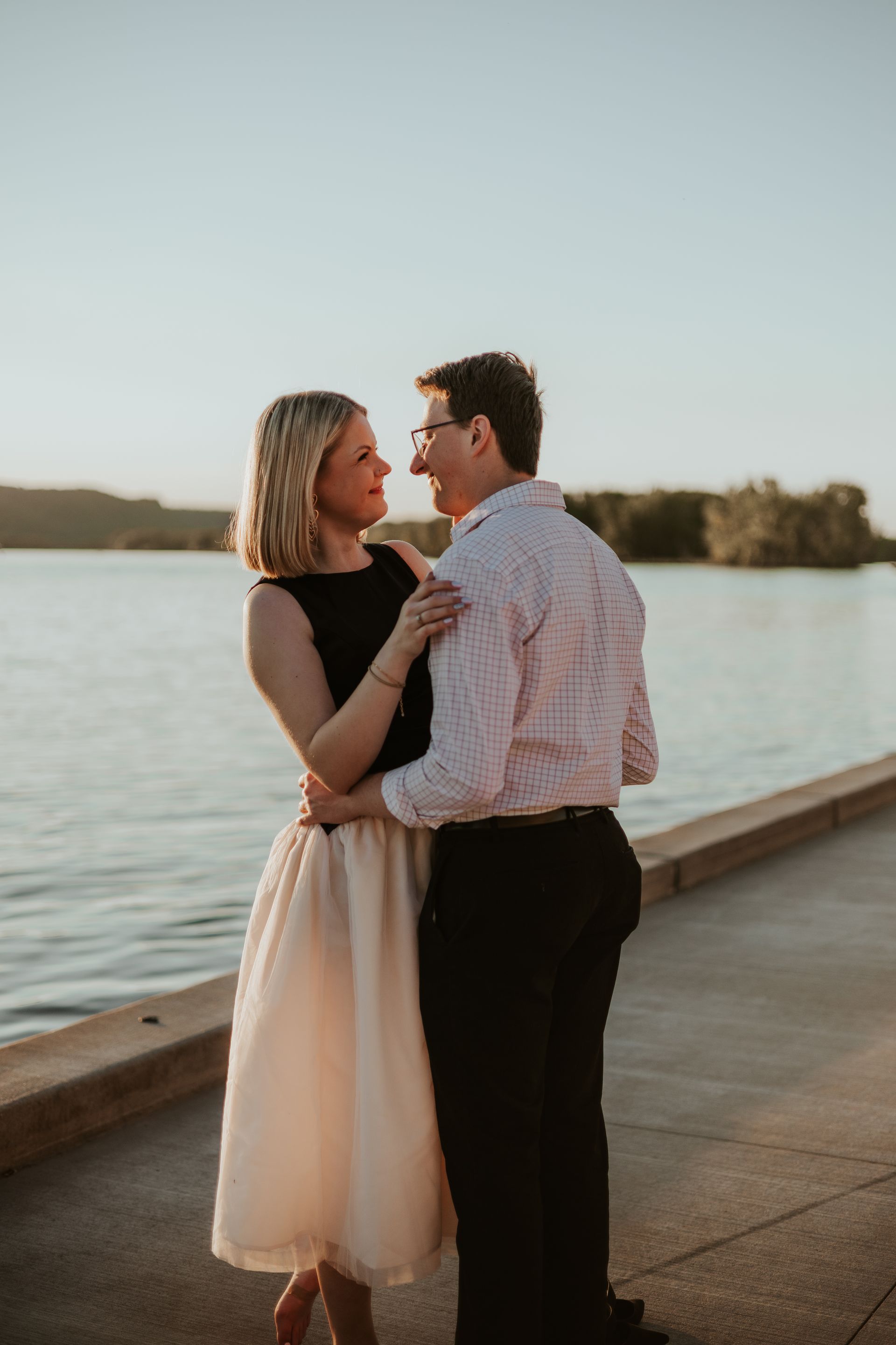 Couple standing close on a waterfront boardwalk at sunset, facing each other by the water.