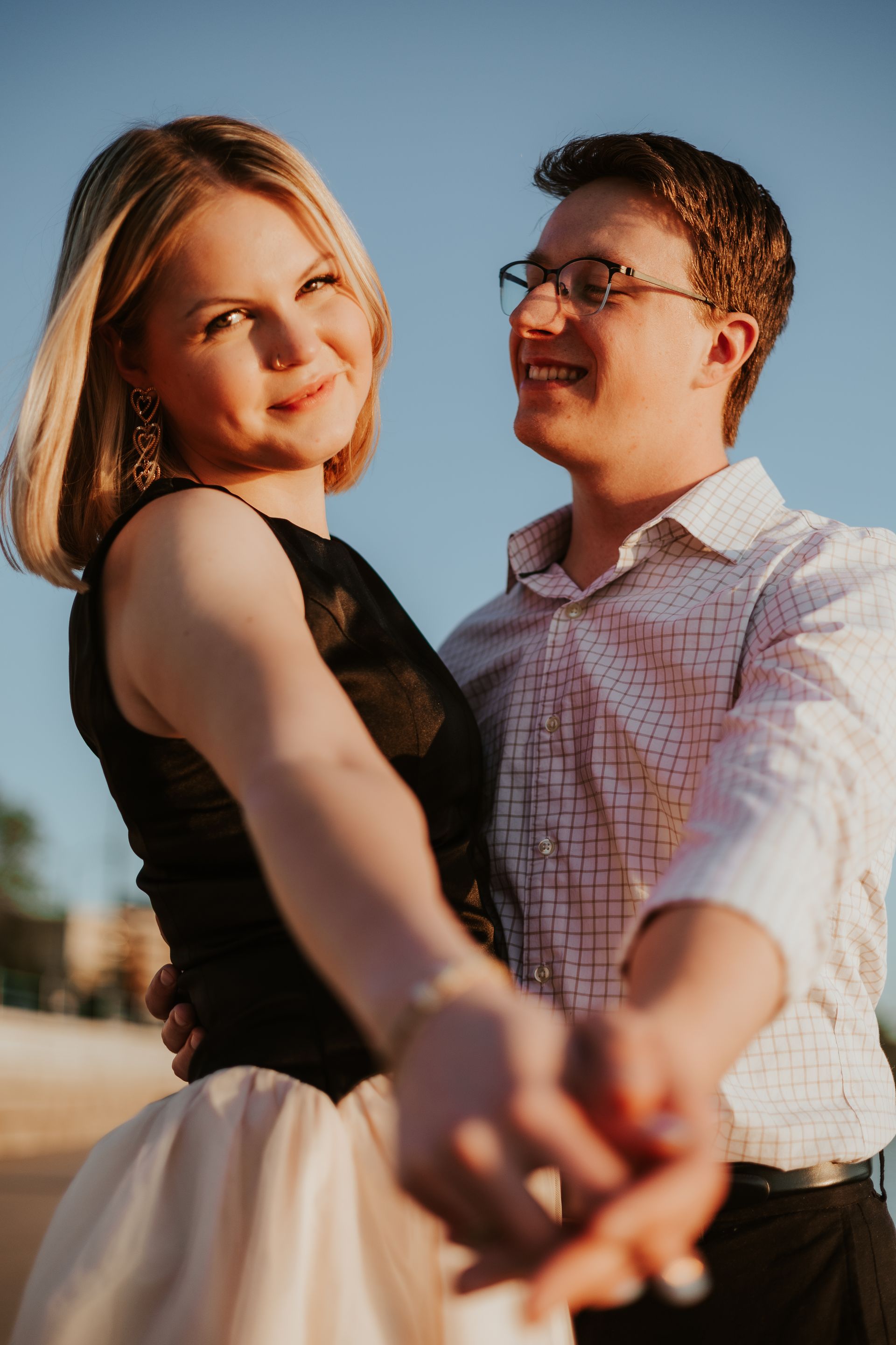 Smiling couple holding hands outdoors at sunset, with the woman in the foreground and the man behind her.