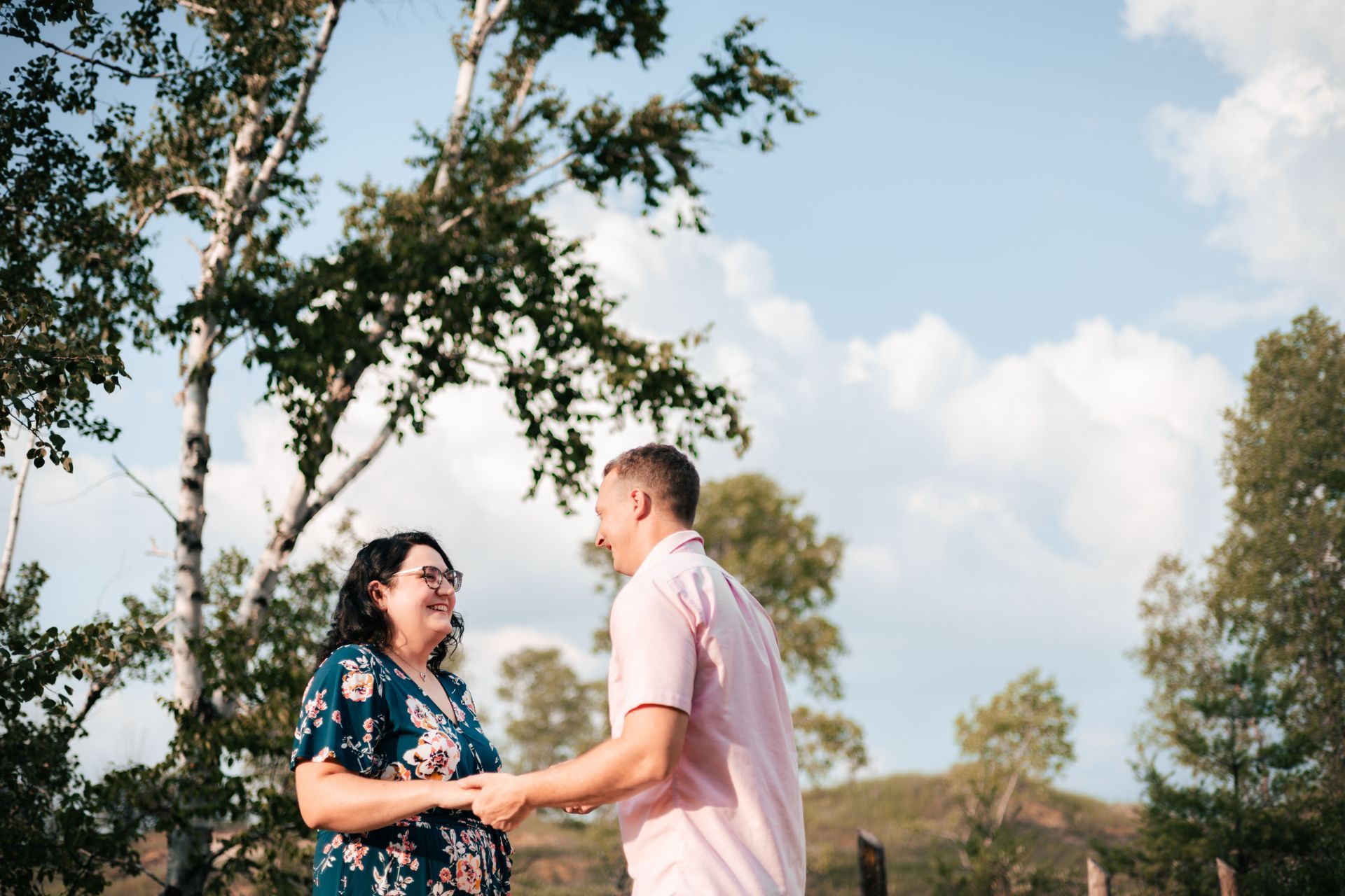 Two people smiling and holding hands outdoors under a tree on a sunny day