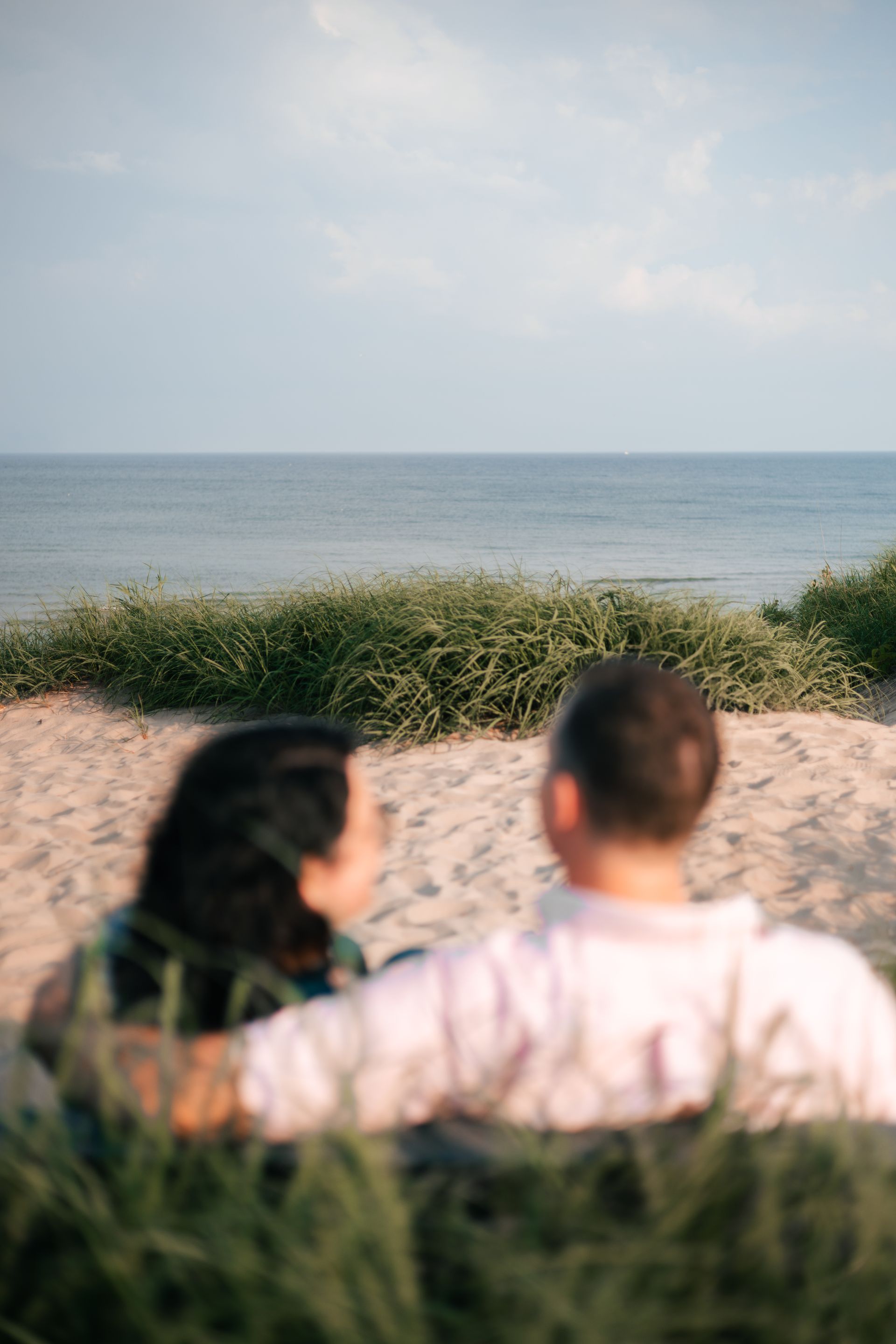 Two people sit on sandy dunes facing a calm ocean under a pale sky