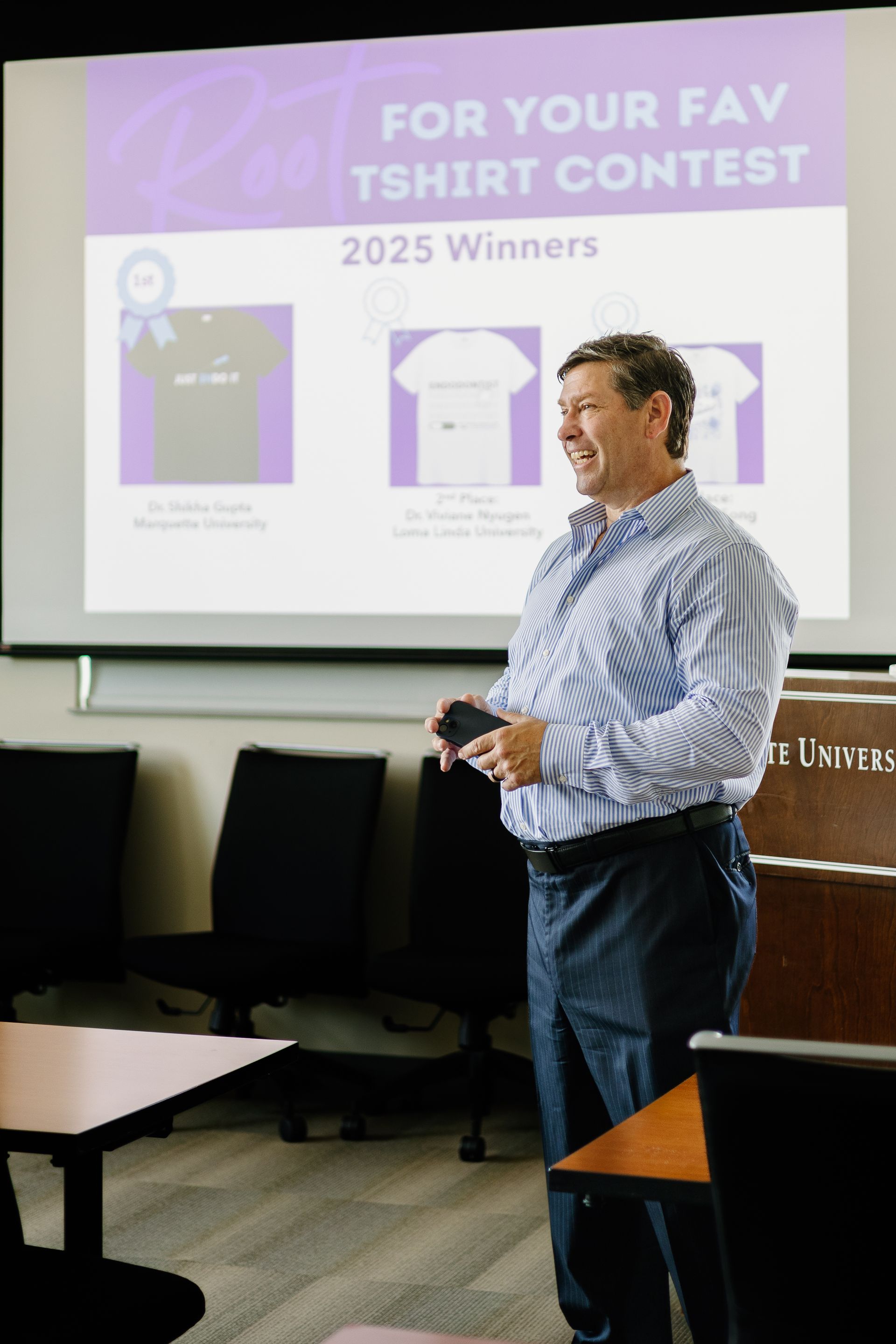 Presenter stands beside a slide reading “For Your Fav T-shirt Contest 2025 Winner” in a meeting room.