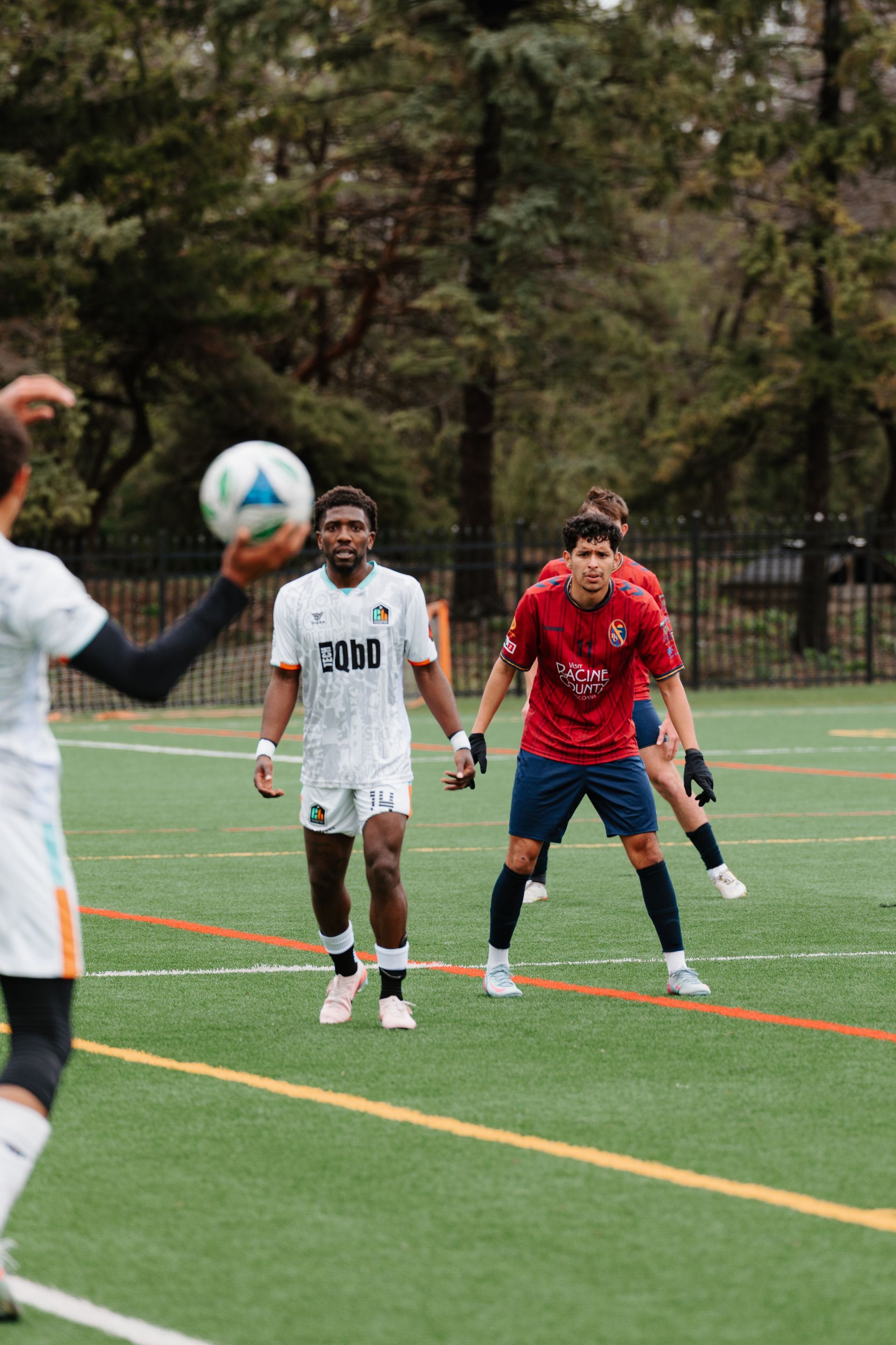 Soccer players on an outdoor turf field, with one player kicking the ball toward two defenders.