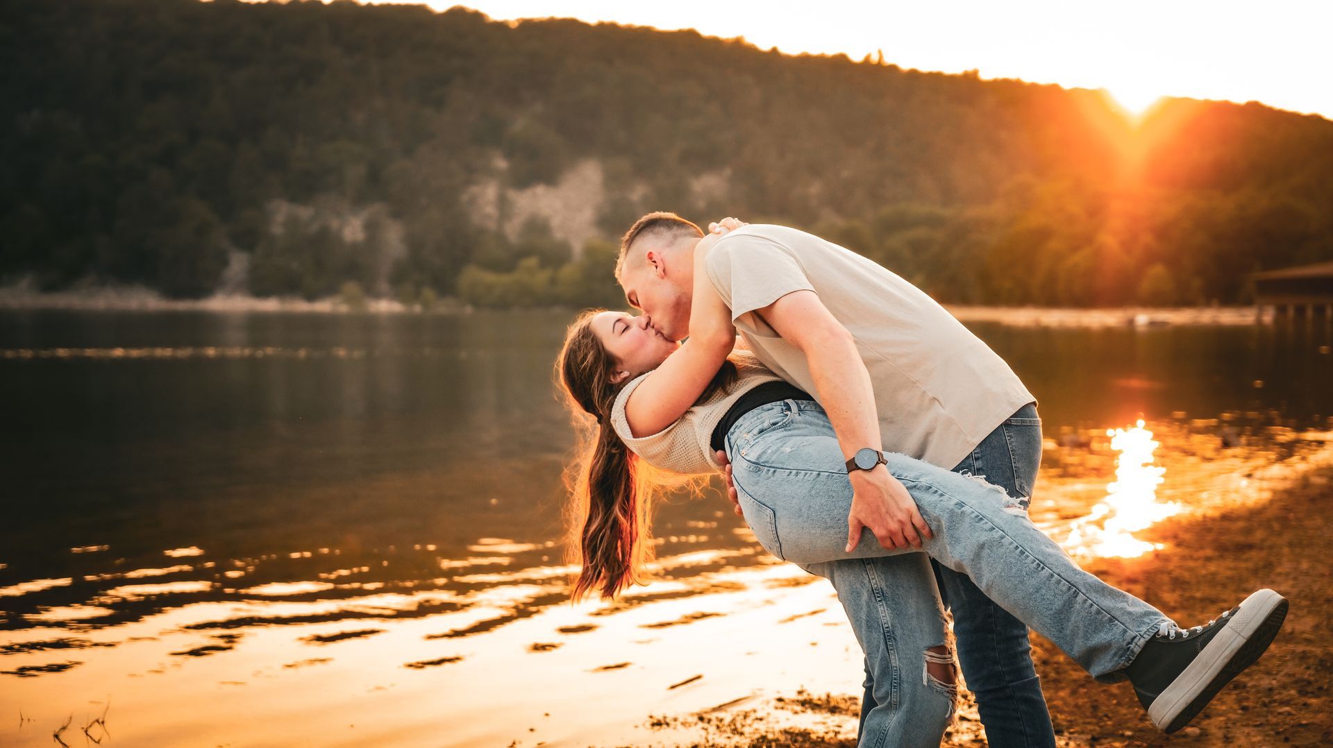 Couple kissing by a lake at sunset, with warm golden light and forested hills in the background
