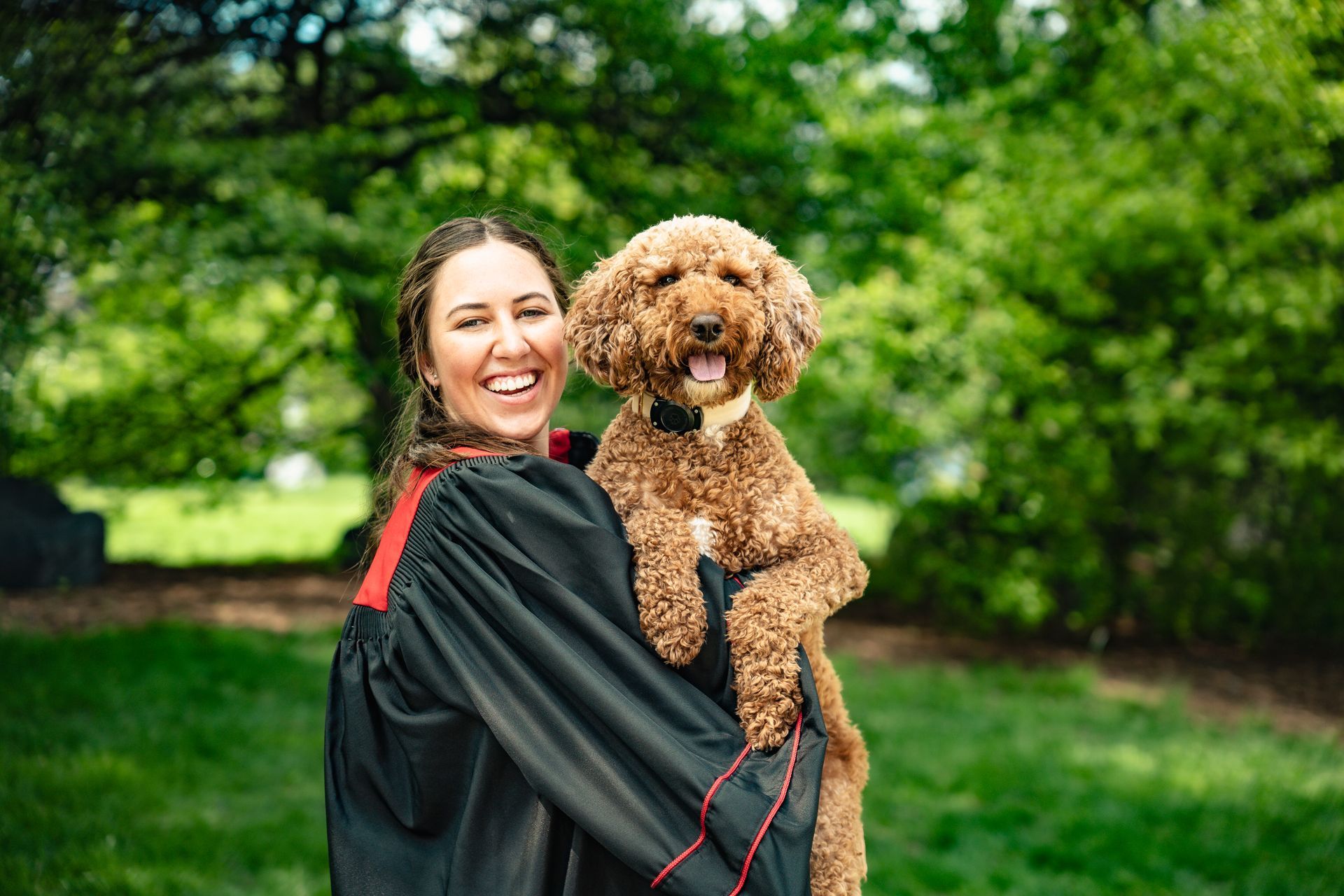 Woman in a black gown holding a curly brown dog outdoors in a green park, both smiling.