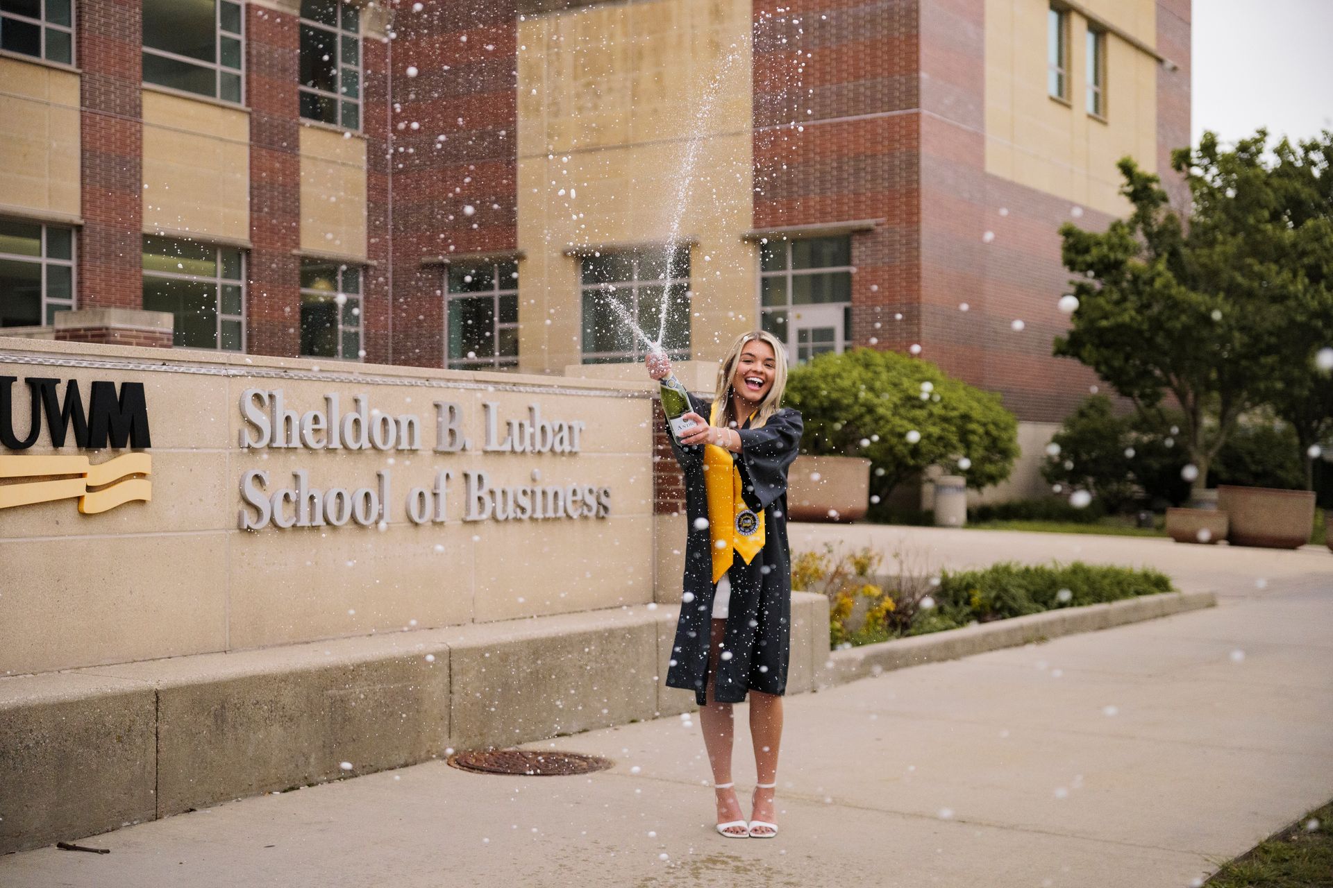 Person in graduation gown standing outside Sheldon B. Luman School of Business with fountain and confetti