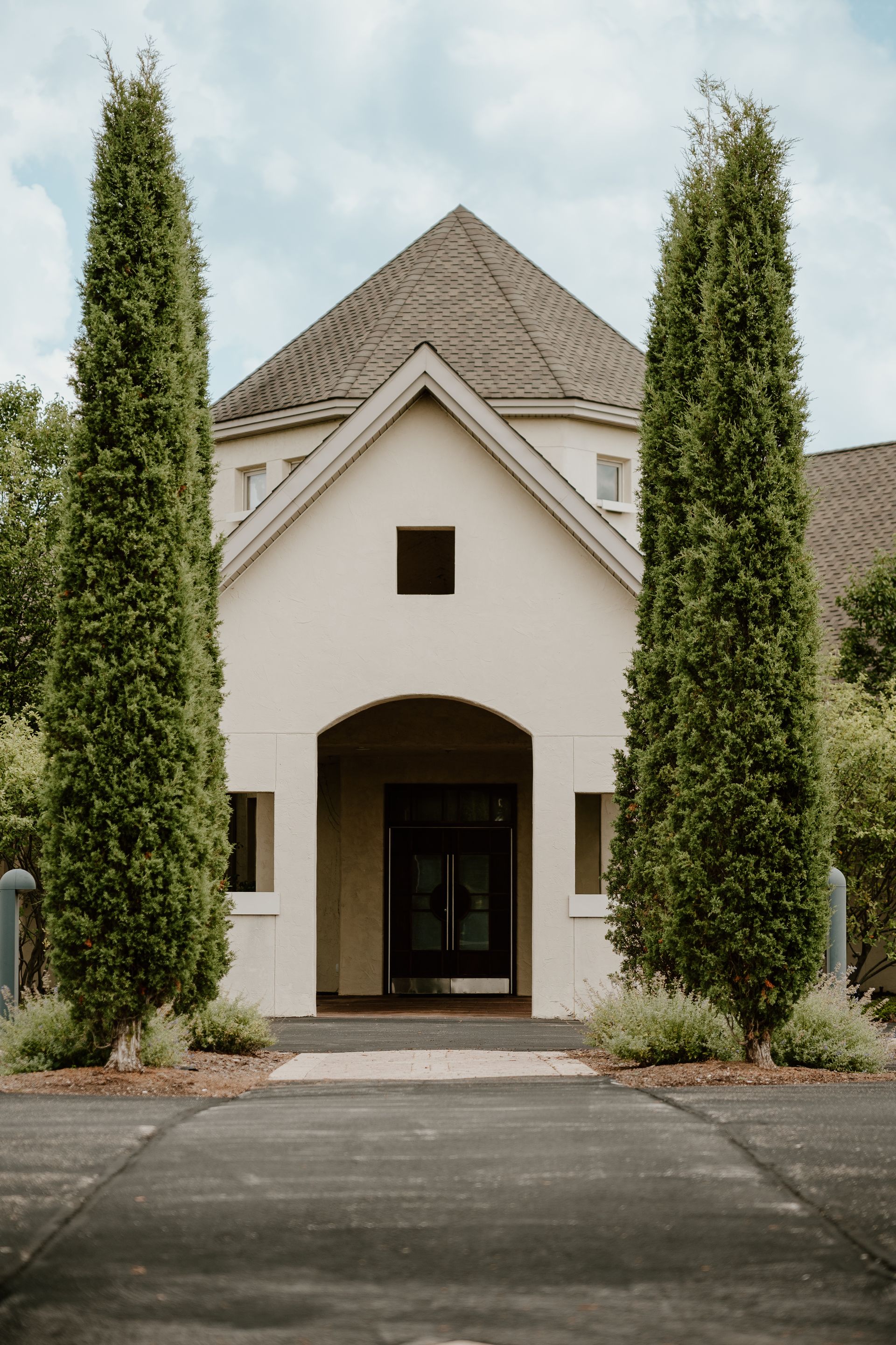 Front view of a white chapel with tall cypress trees and a tiled roof, centered on a paved driveway