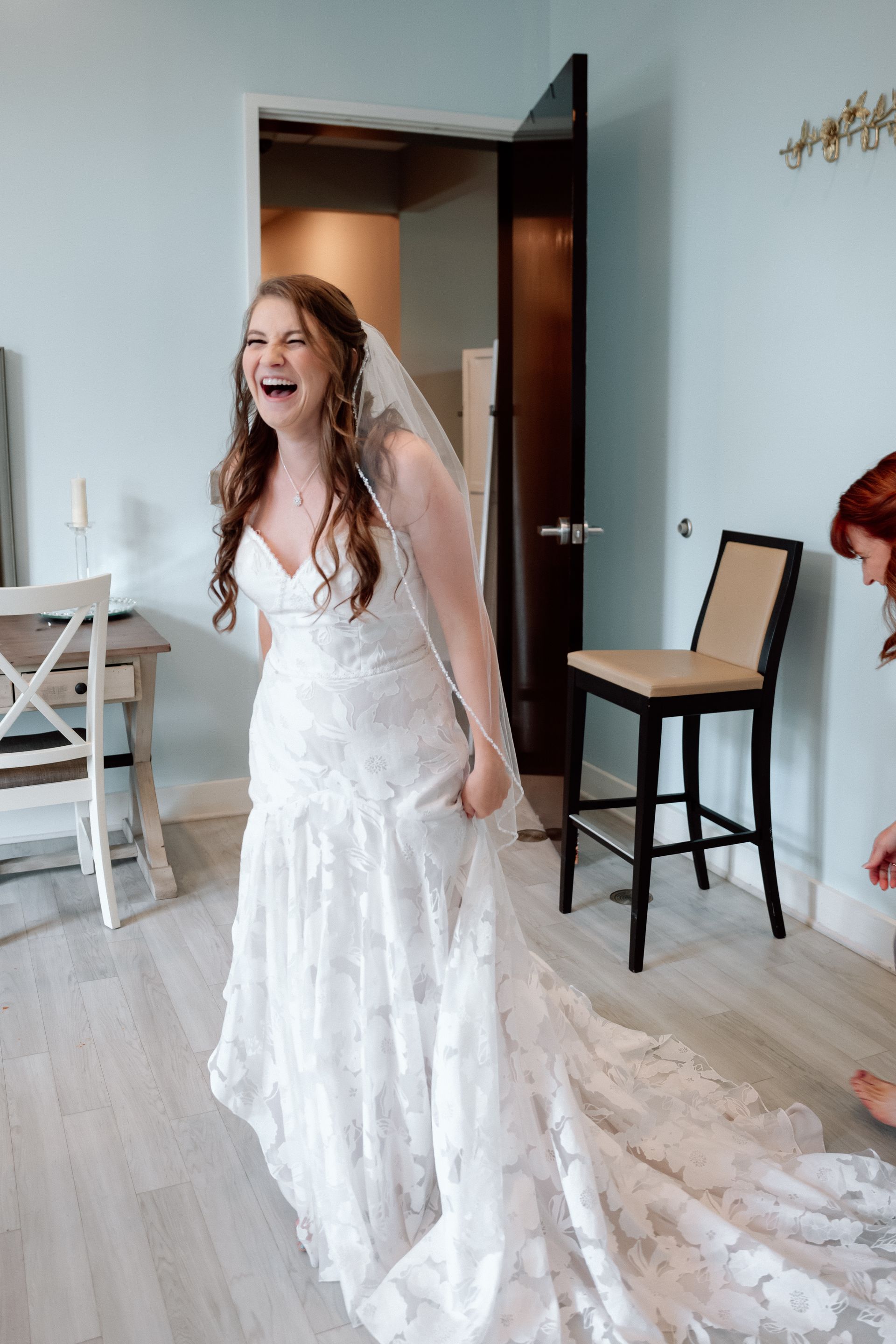 Bride in a white wedding gown laughing in a room with open door and chair nearby