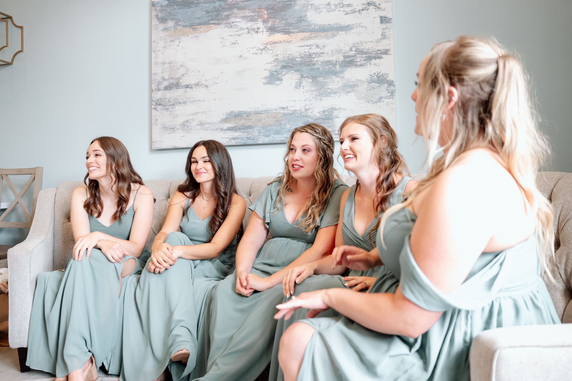 Five women in matching mint dresses sit on sofas chatting in a bright living room.