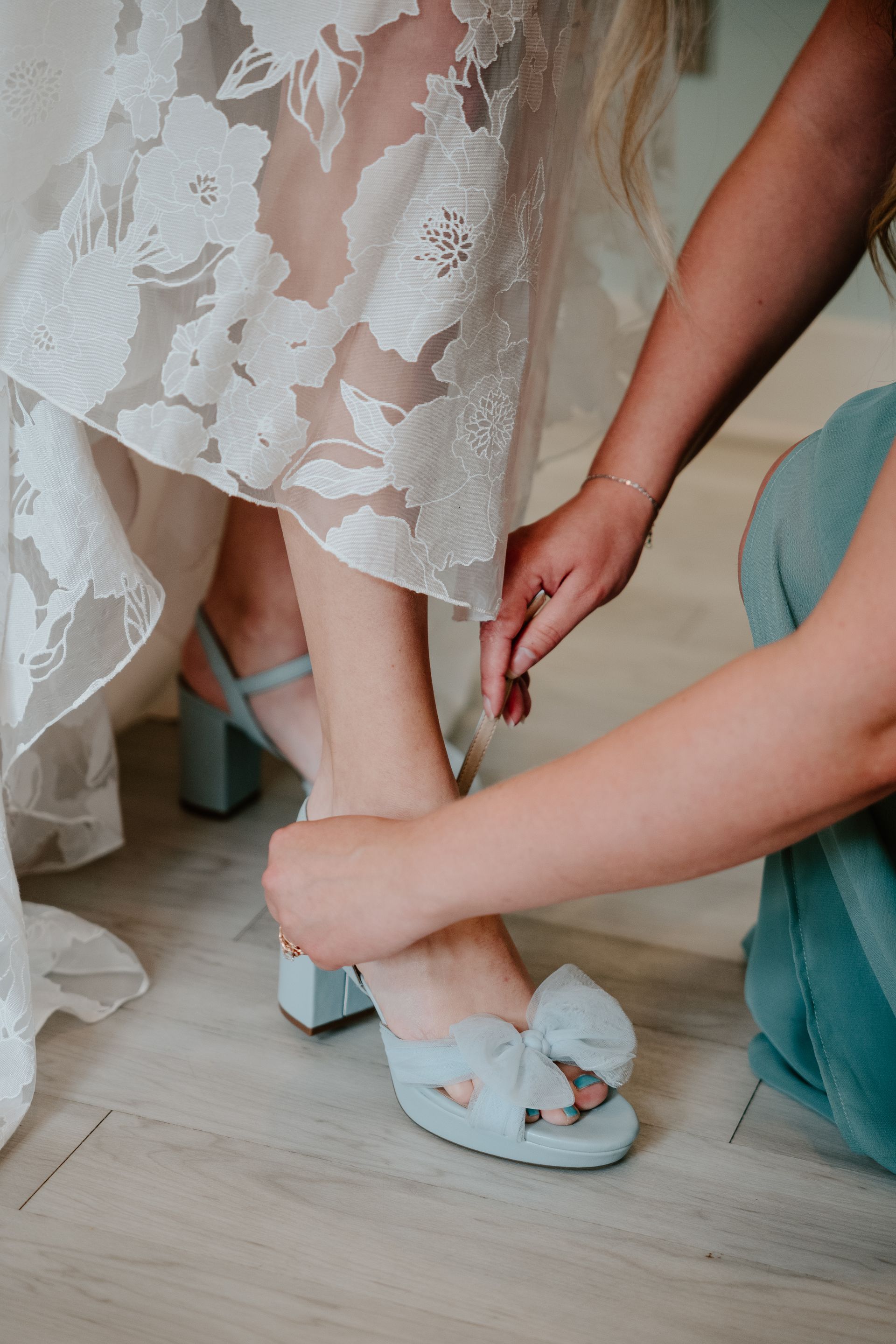 Person adjusting light blue shoe strap on another person's foot beside a white lace dress