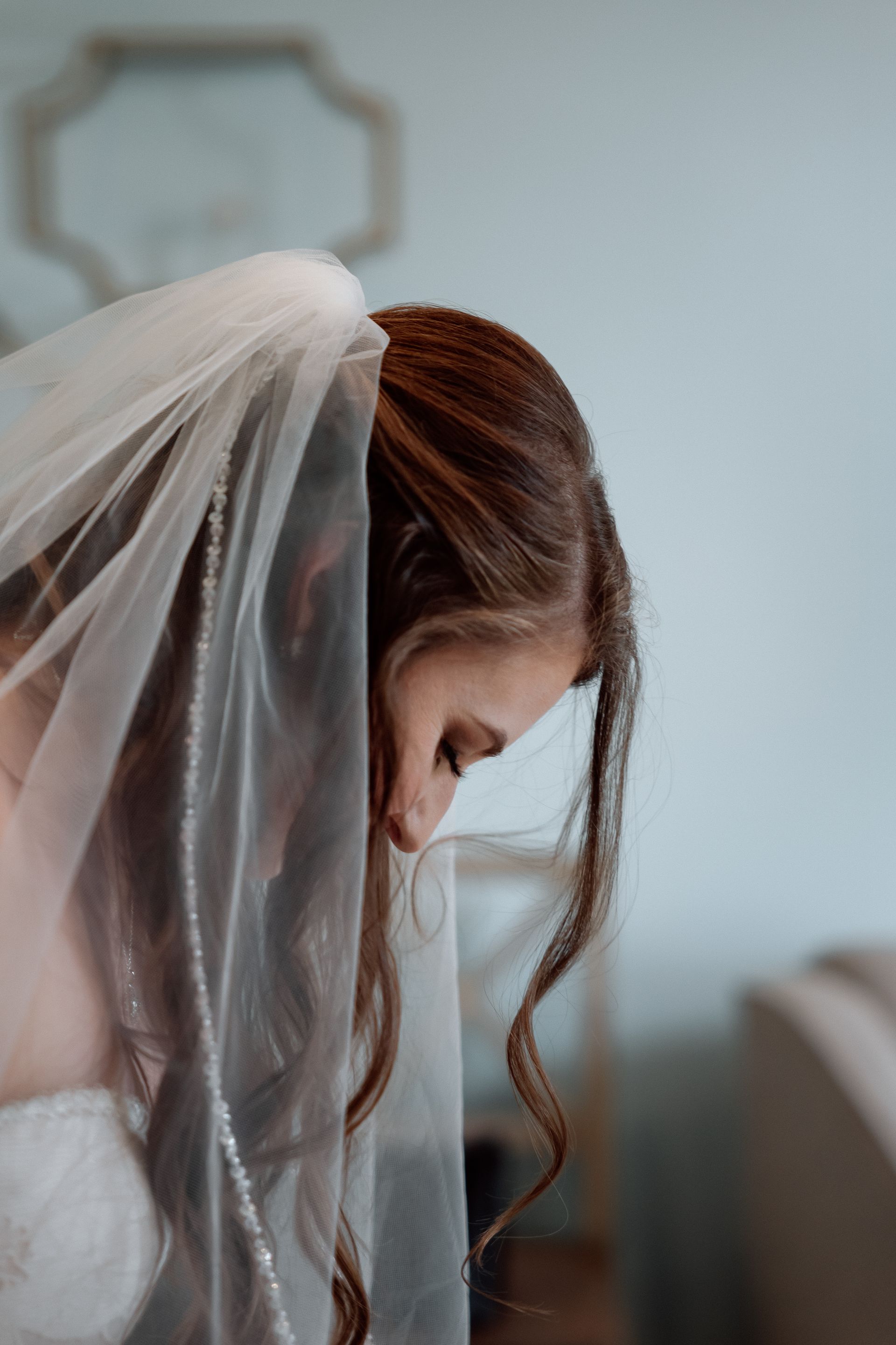 Bride adjusting her veil, with long brown hair, indoors by a light wall