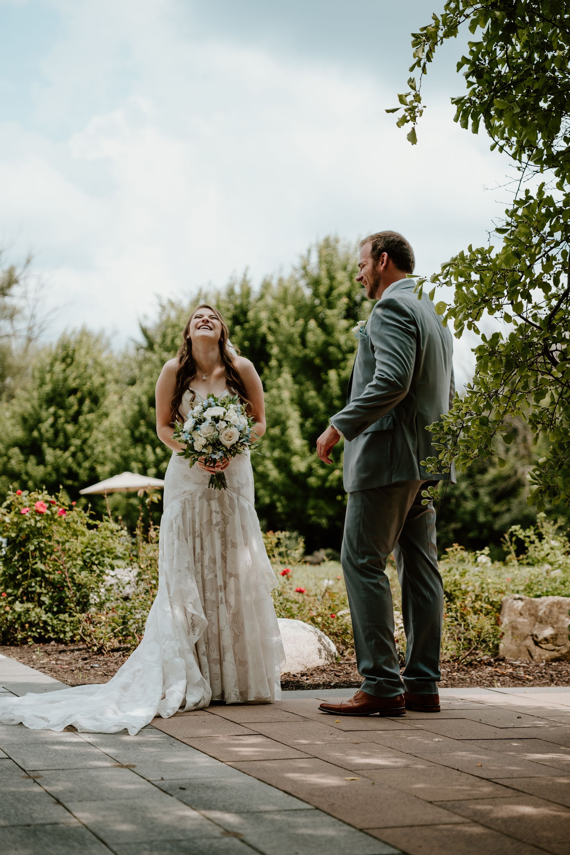 Bride in a white lace dress holding a bouquet faces a man in a suit in a garden setting.