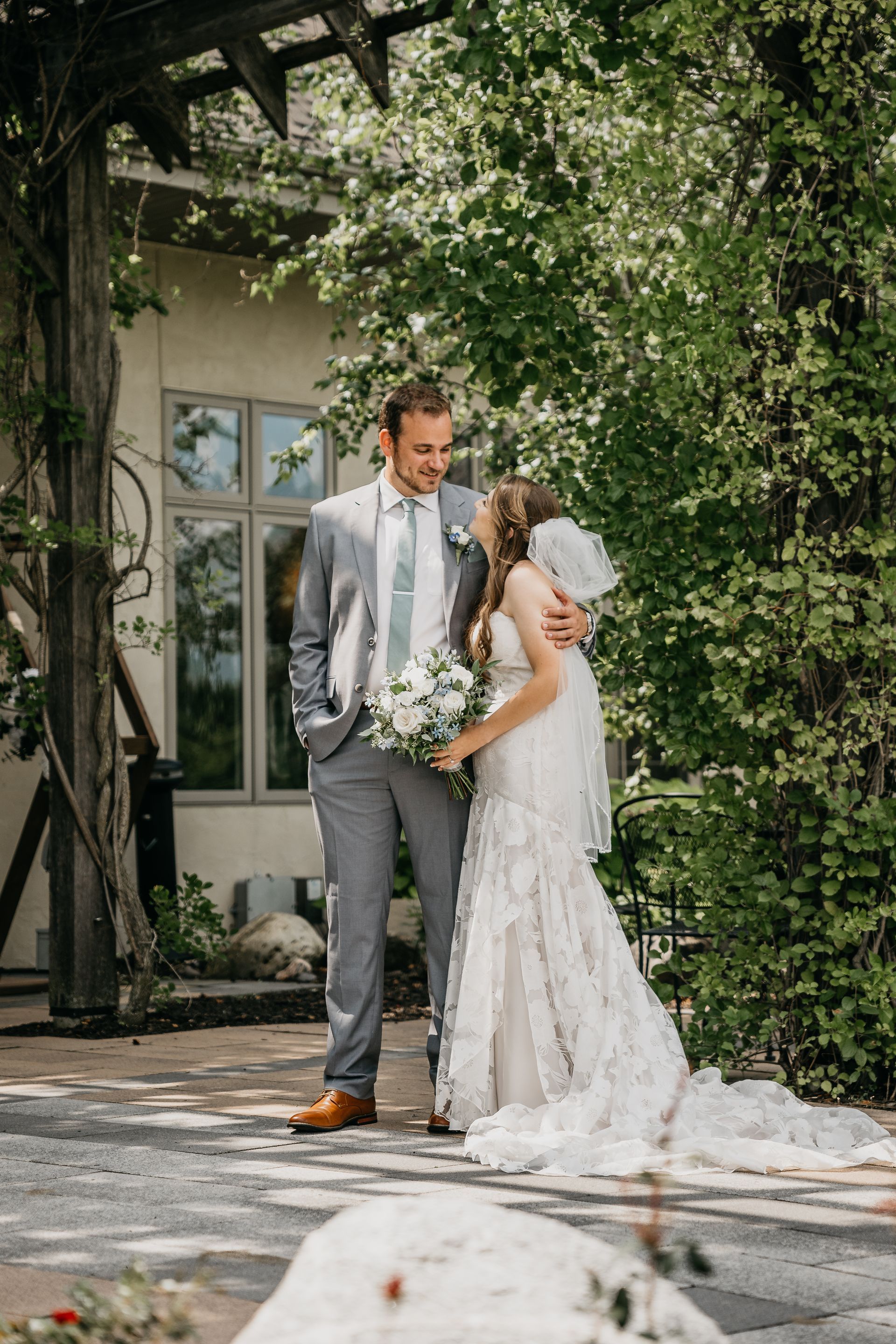 Bride and groom standing together under greenery at an outdoor wedding, holding a bouquet