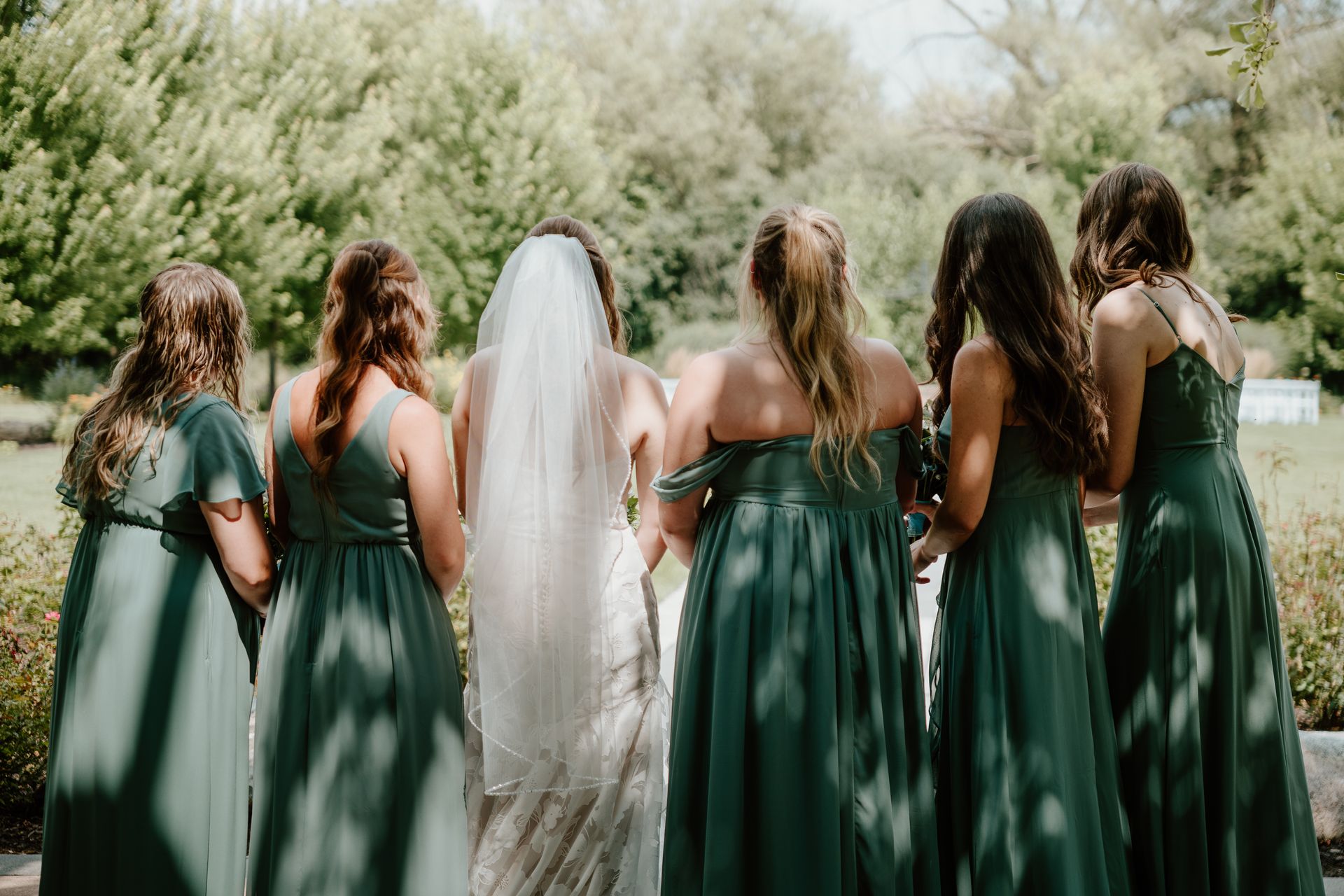 Six people in green dresses and one in a white veil stand outdoors with their backs to the camera.