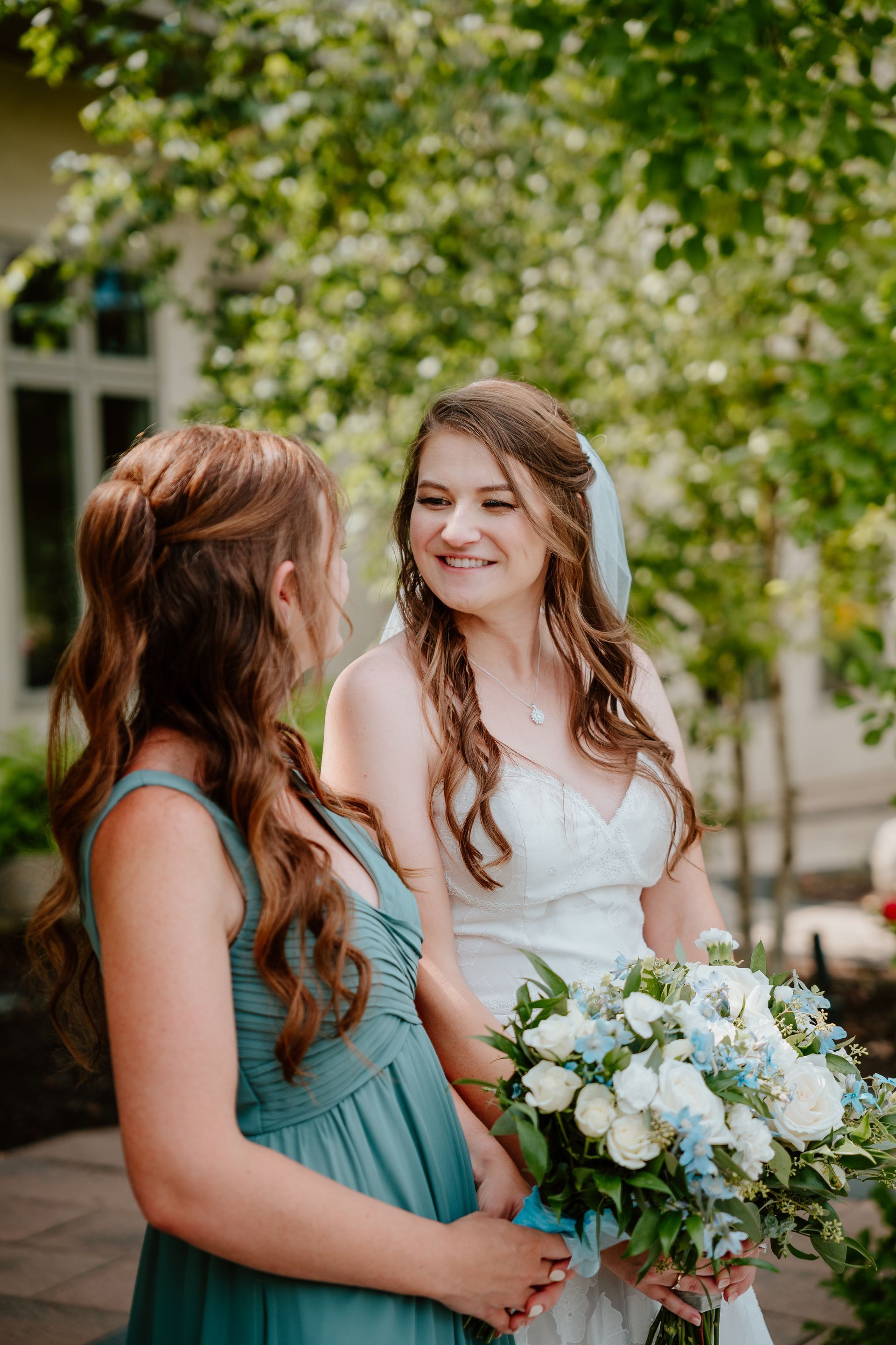 Two women in pastel dresses smiling outdoors, one holding a white and blue bouquet, in a garden setting
