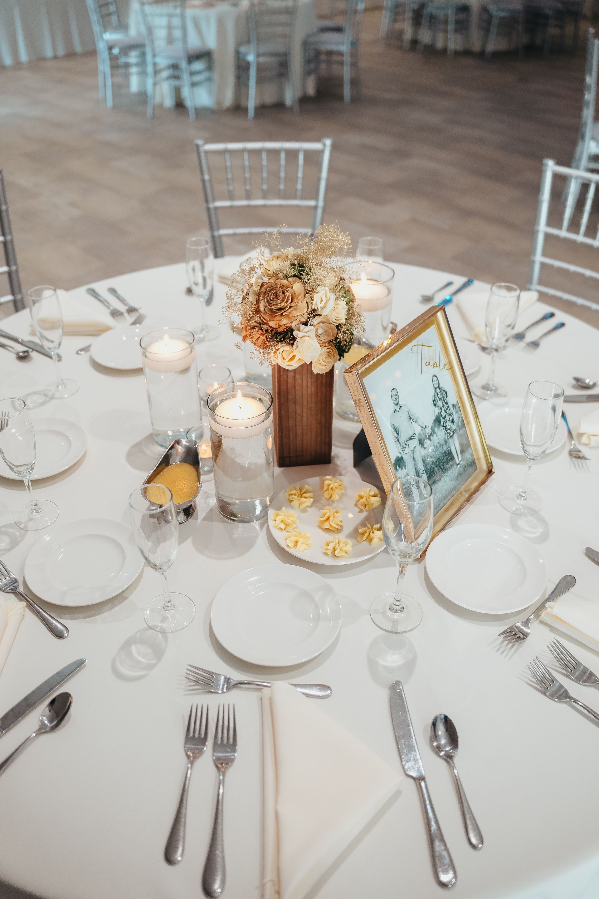 Round banquet table set with white plates, silverware, candles, and a floral centerpiece on a white chair-backed floor.