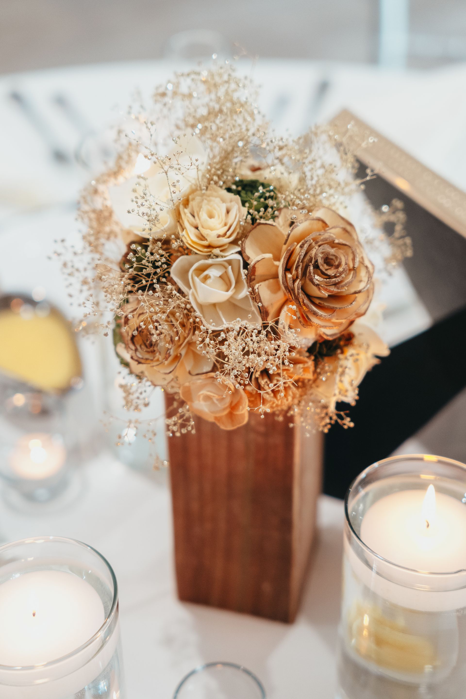 Cream and tan floral centerpiece in a wooden vase with candles on a softly lit table