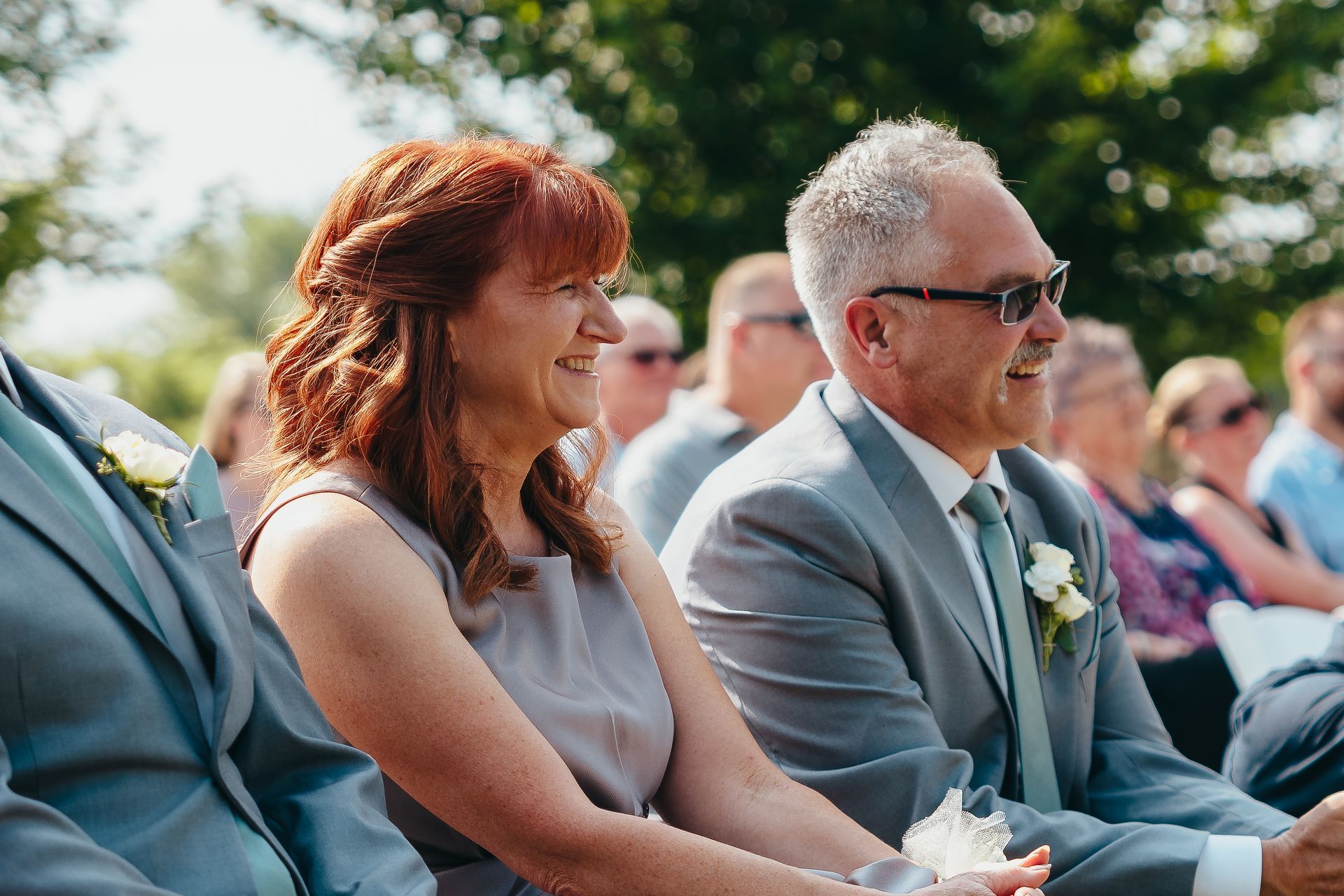 Two seated guests smiling outdoors at a sunny ceremony, one in a light gray dress and one in a gray suit and sunglasses