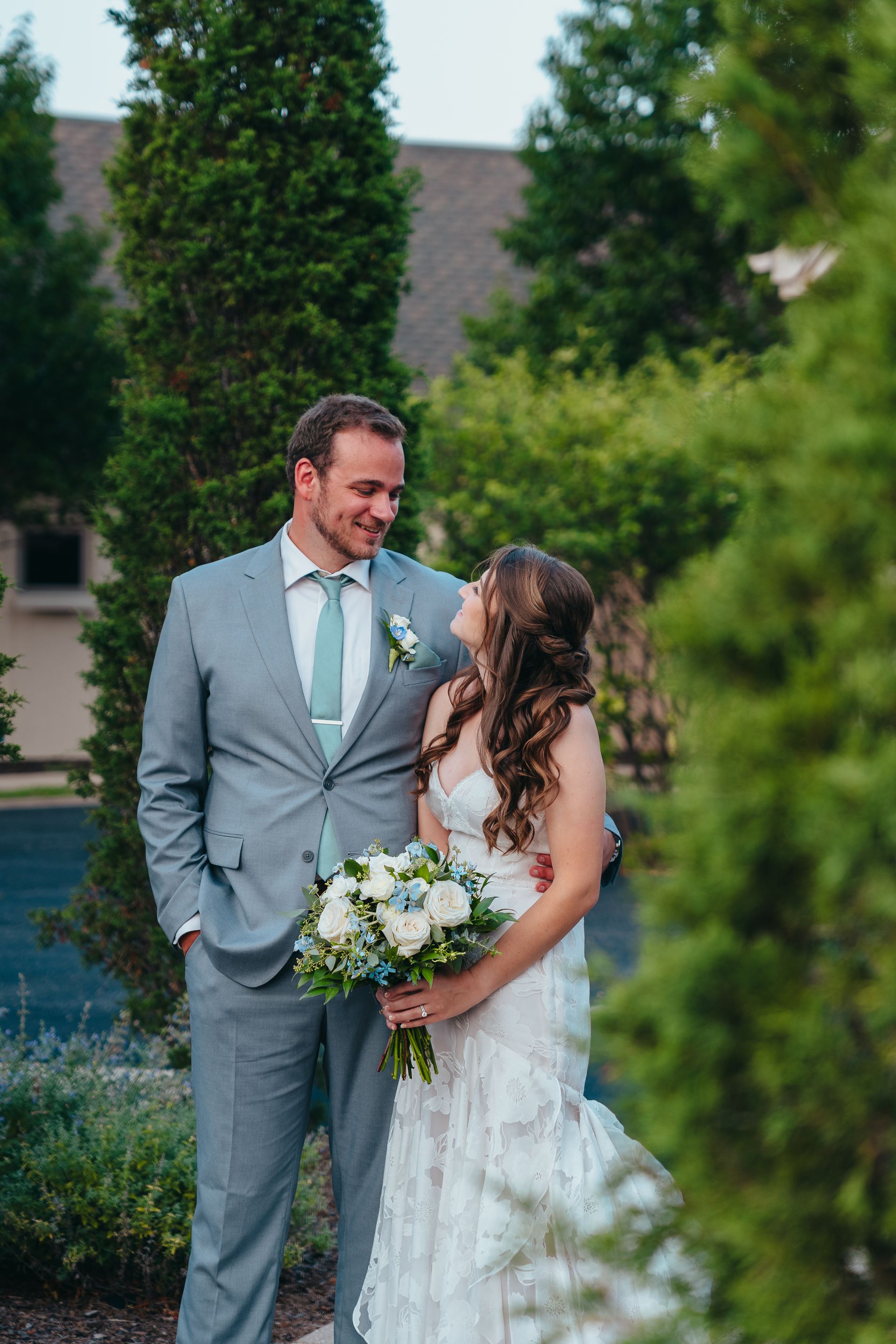 Wedding couple smiling outdoors in front of greenery, bride holding bouquet and looking up at groom in a gray suit