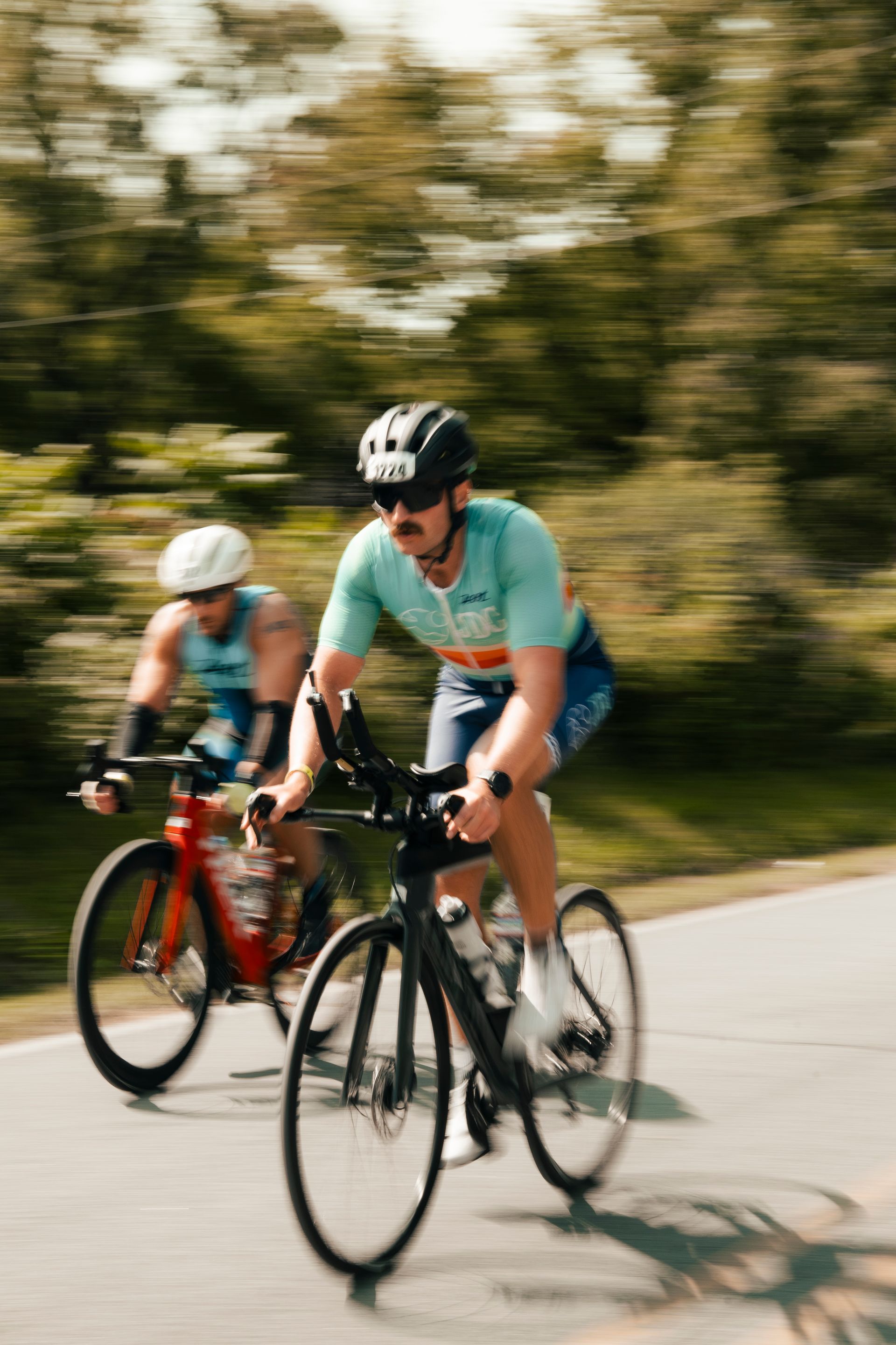 Two cyclists riding on a road, one in a teal shirt leading, with blurred trees in the background