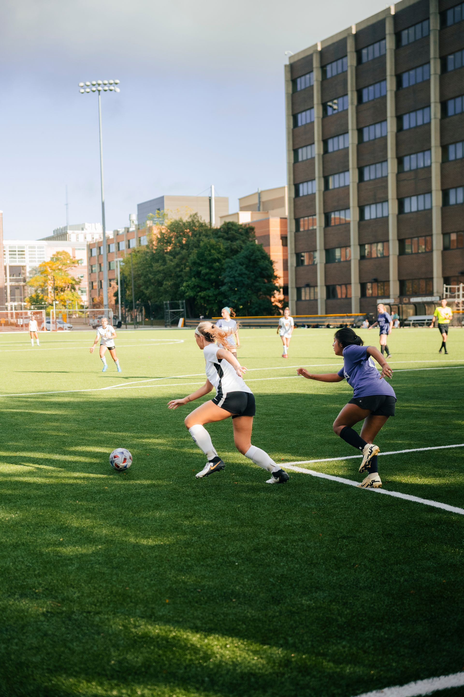 Soccer players contesting the ball on a sunny field near campus buildings