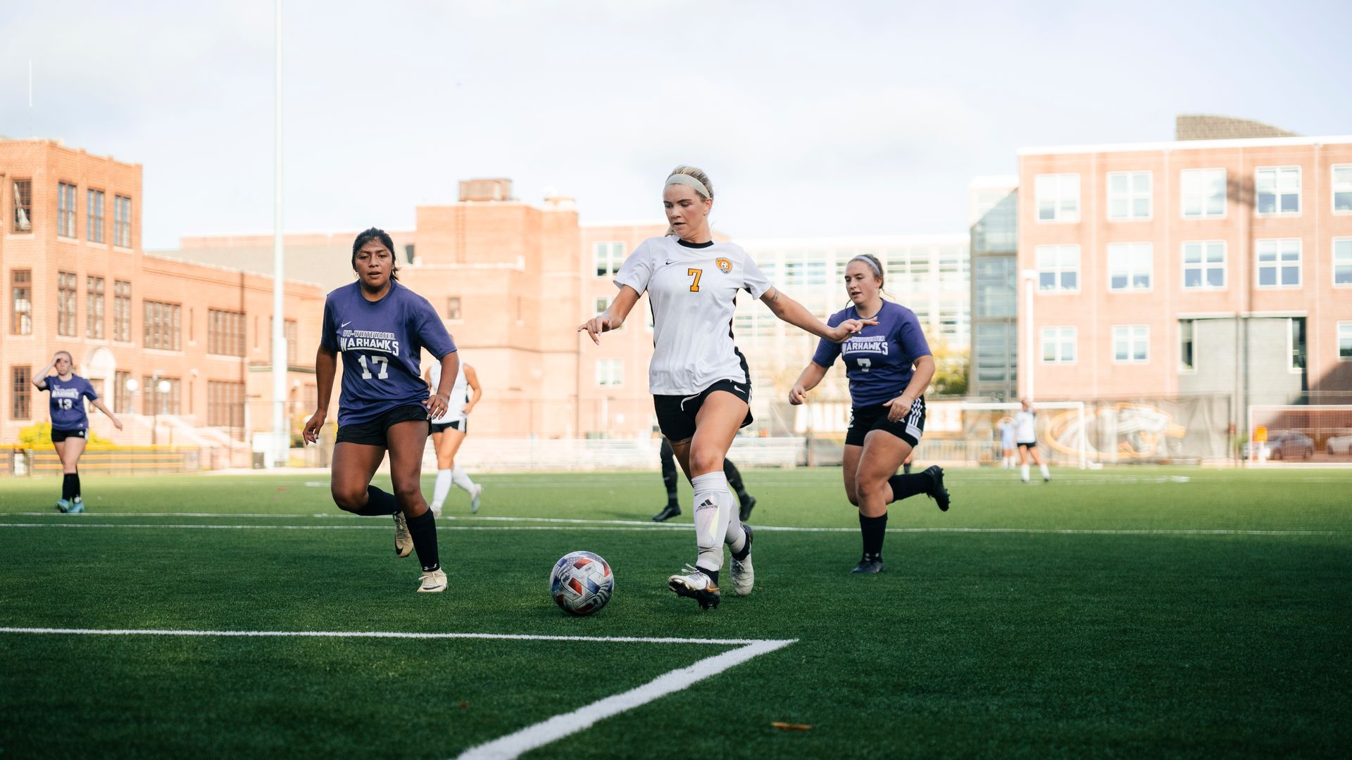 Soccer players sprint toward the ball on a sunny field in front of campus buildings.