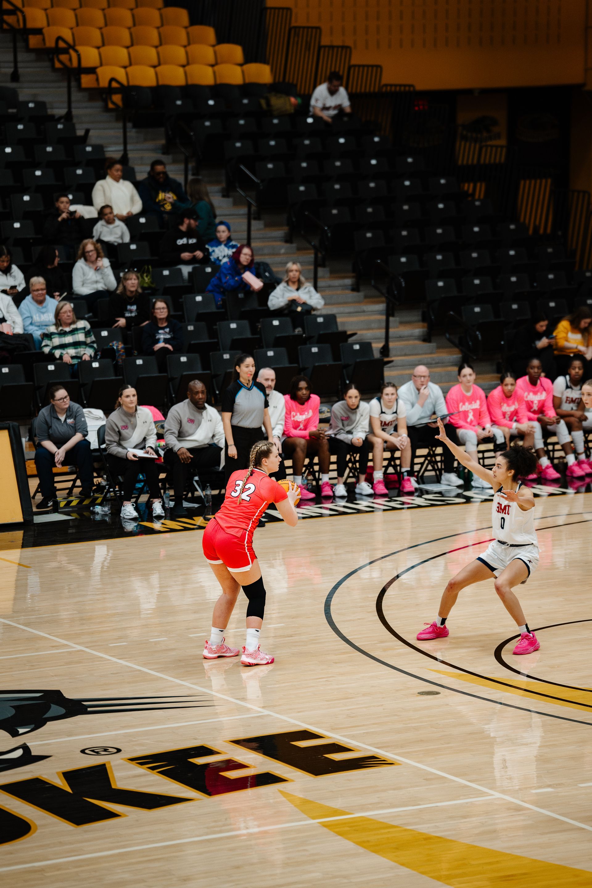 Women’s basketball game with orange and white jerseys on a hardwood court, bench and spectators in the background