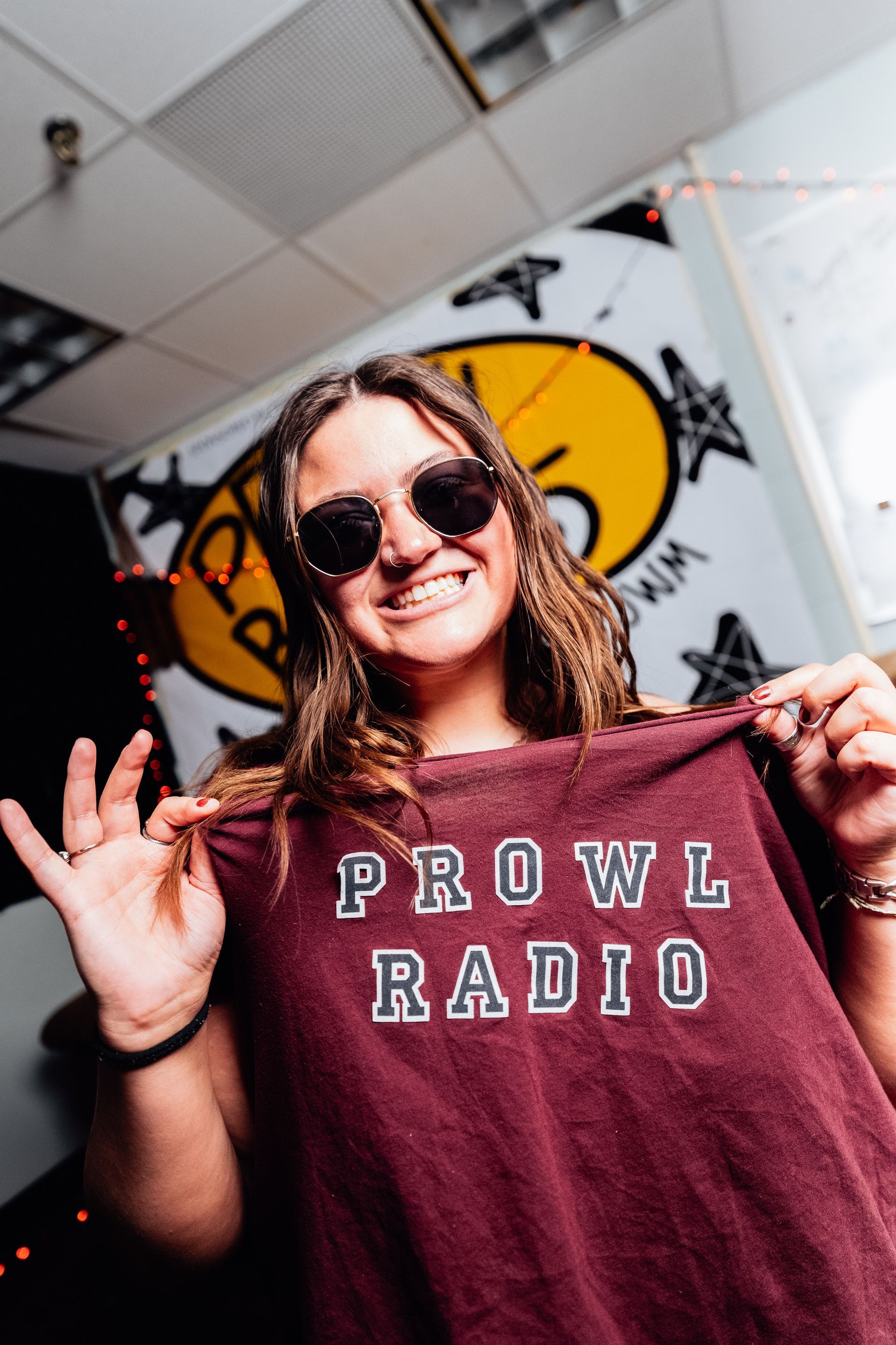 Smiling woman in sunglasses holding up a maroon “PROWL RADIO” shirt
