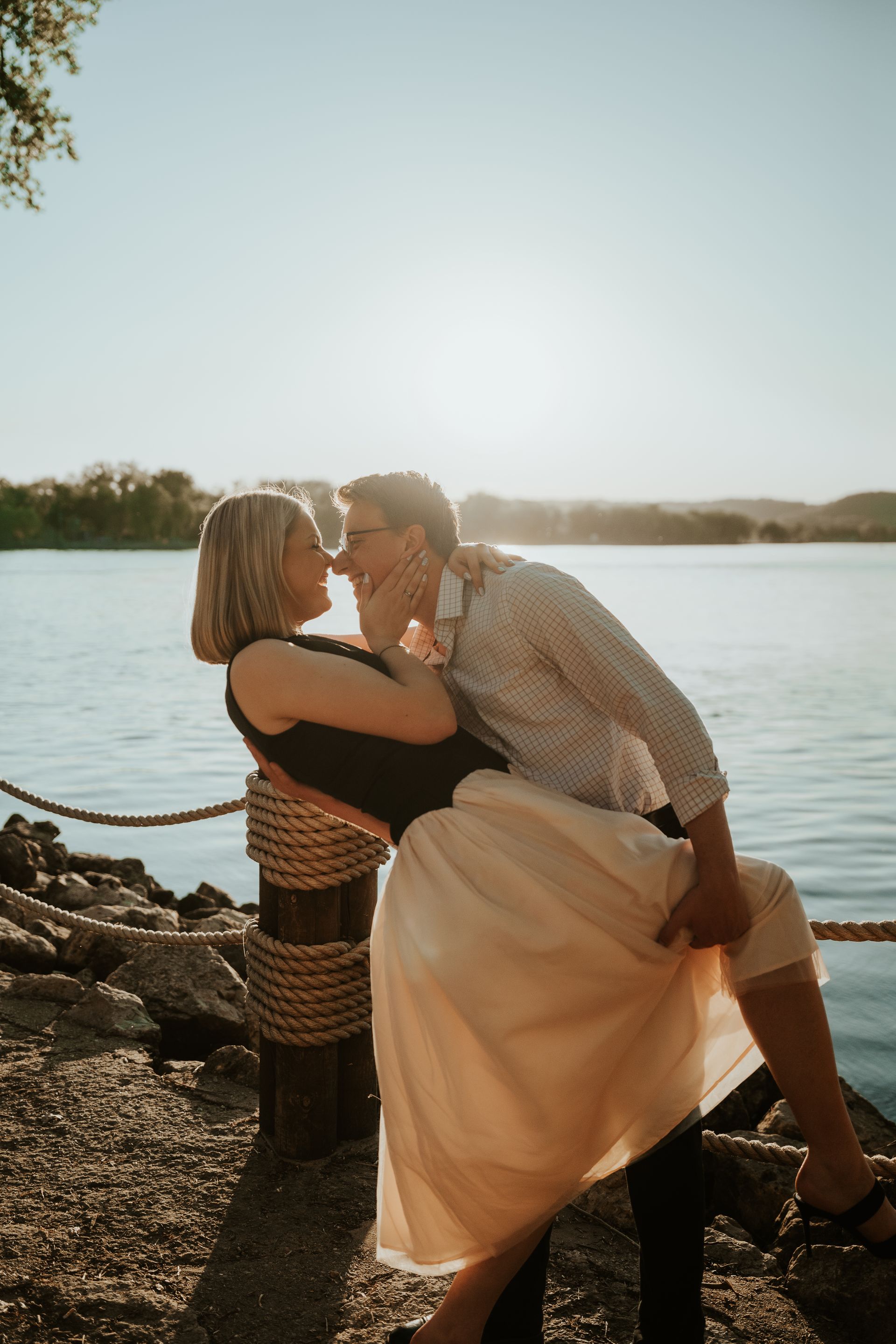 Two people kissing by a rocky shoreline, with water and a bright sky in the background.