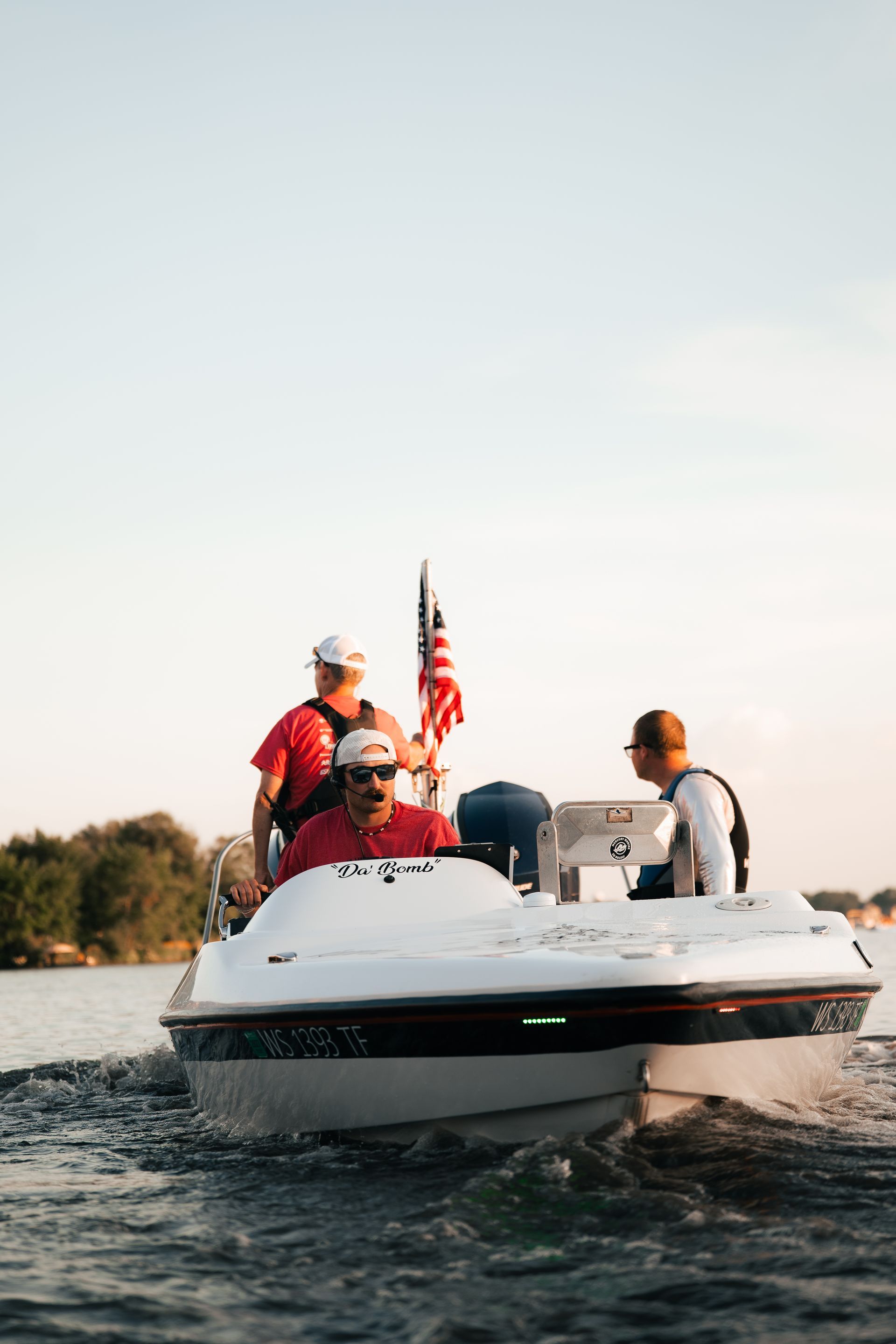 Three people on a white speedboat on calm water, with an American flag behind them.