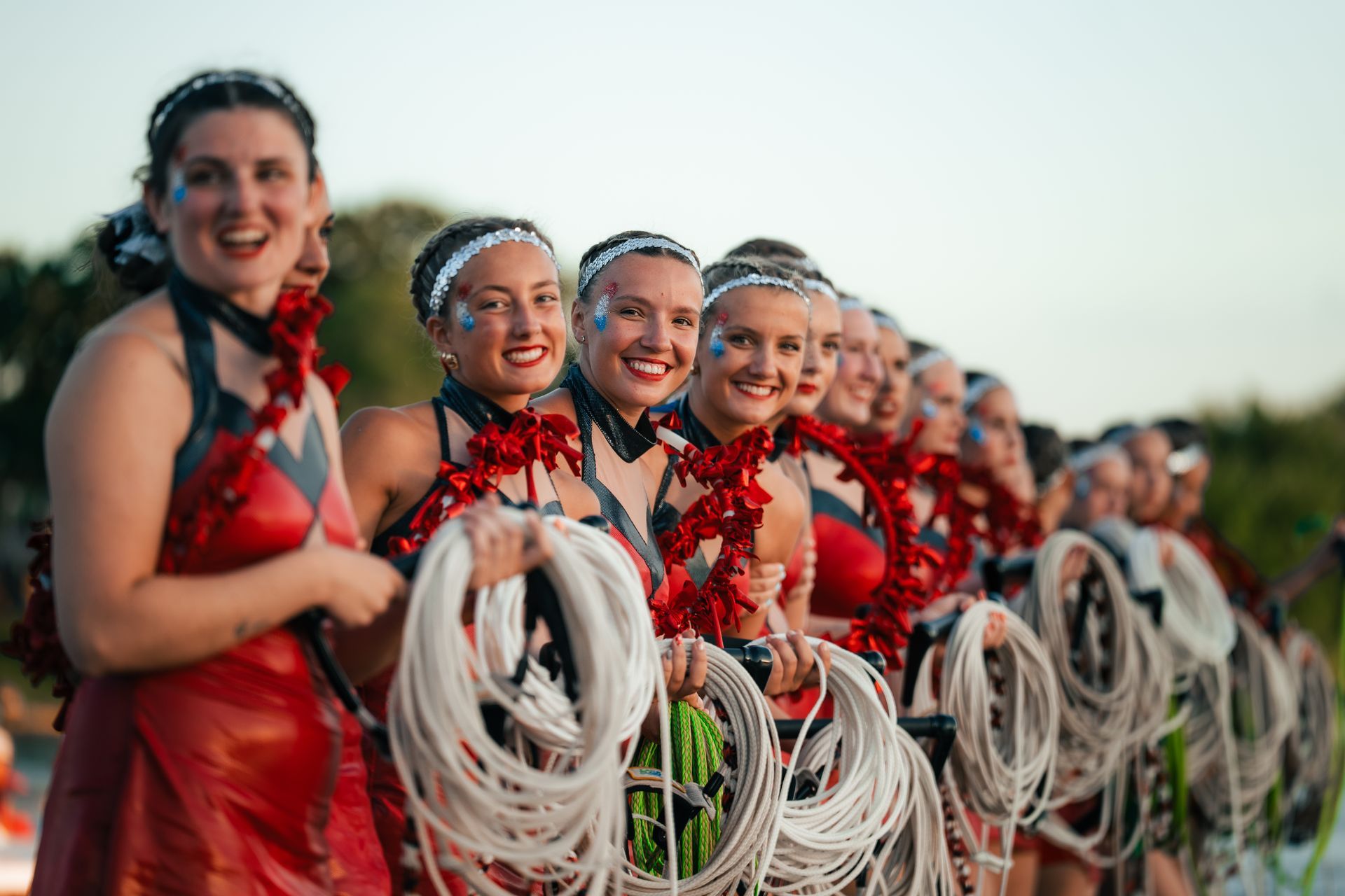 Smiling performers in red costumes hold white ribbon hoops outdoors at sunset