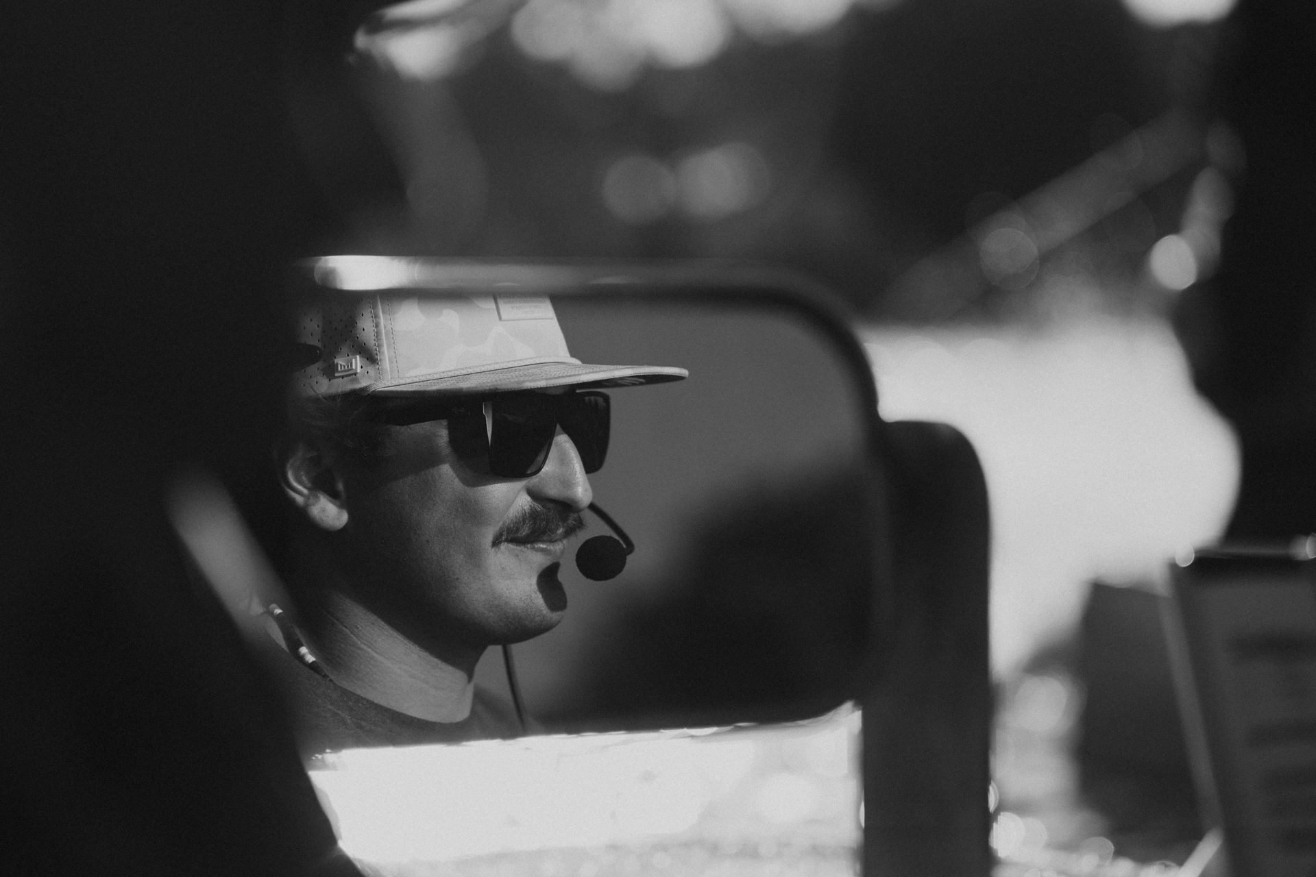 Black-and-white side profile of a man in sunglasses and a hat, seen through a car mirror.