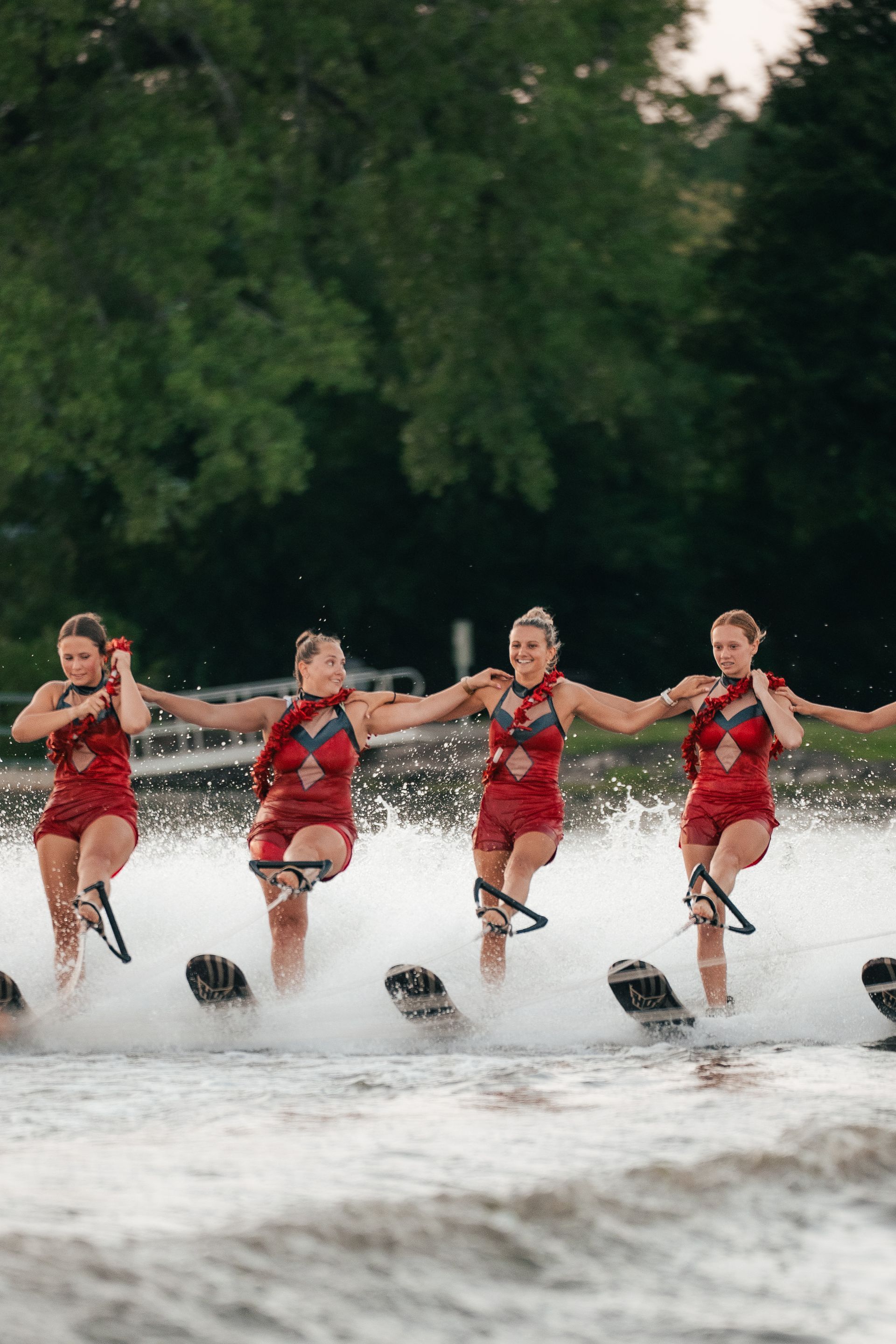 Four synchronized water skiers in red outfits glide across a lake, spraying water in front of a wooded shore.