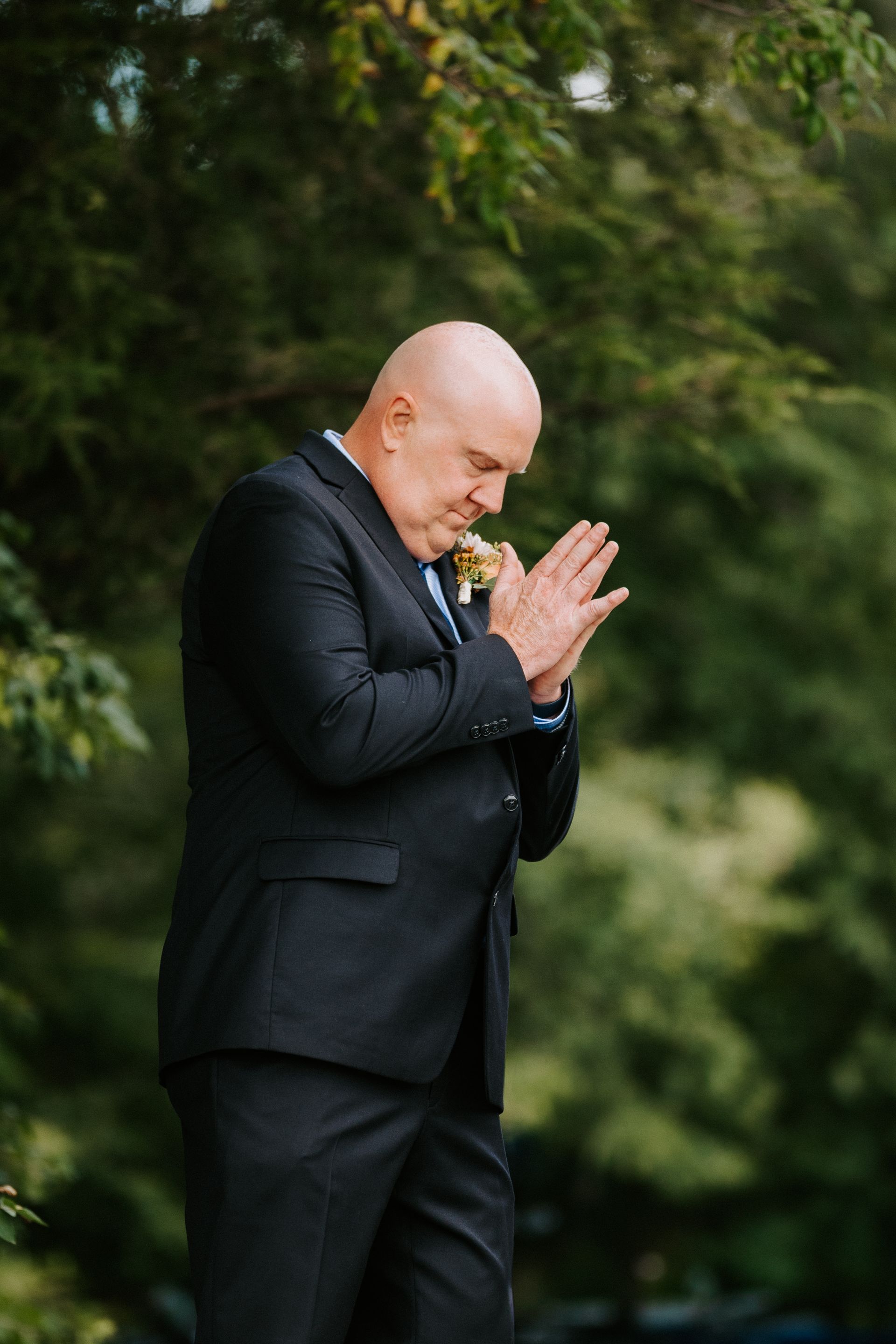 A bald man in a dark suit bows his head with hands clasped outdoors against a blurred green background