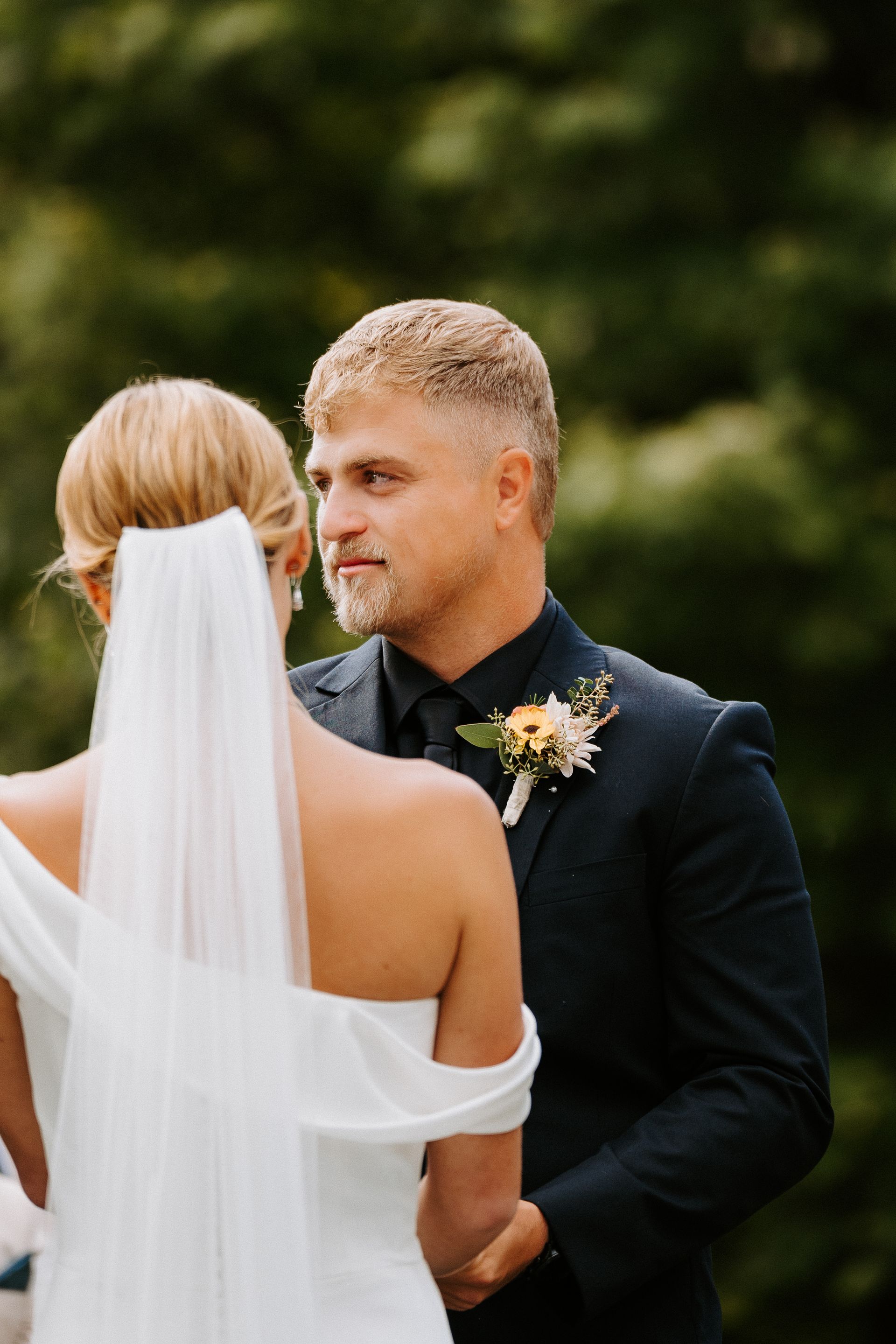 Bride and groom holding hands during an outdoor wedding, with a white veil and dark suit.