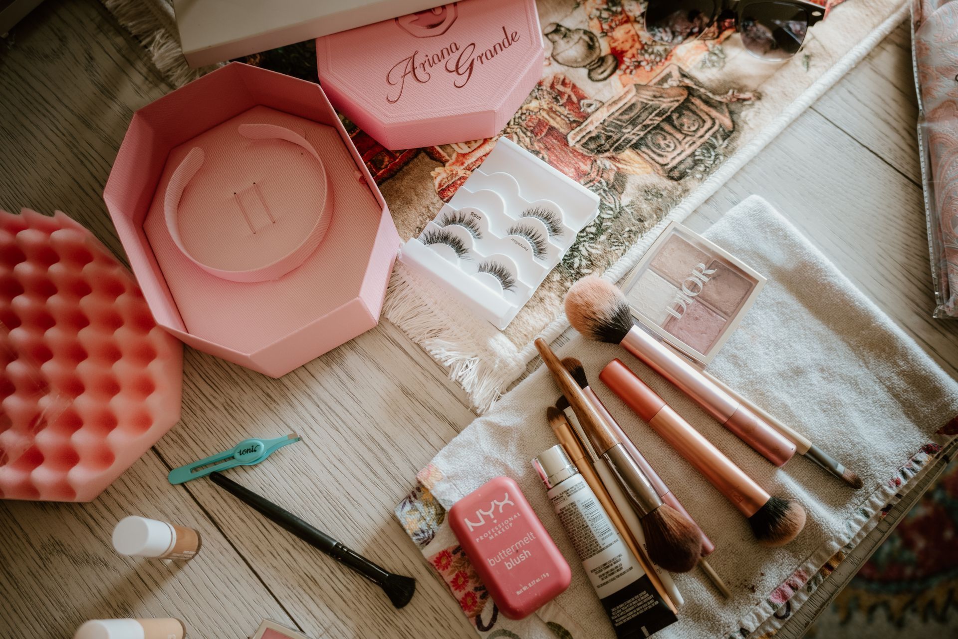 Pink makeup items and brushes arranged on a light wooden table with magazines and a towel