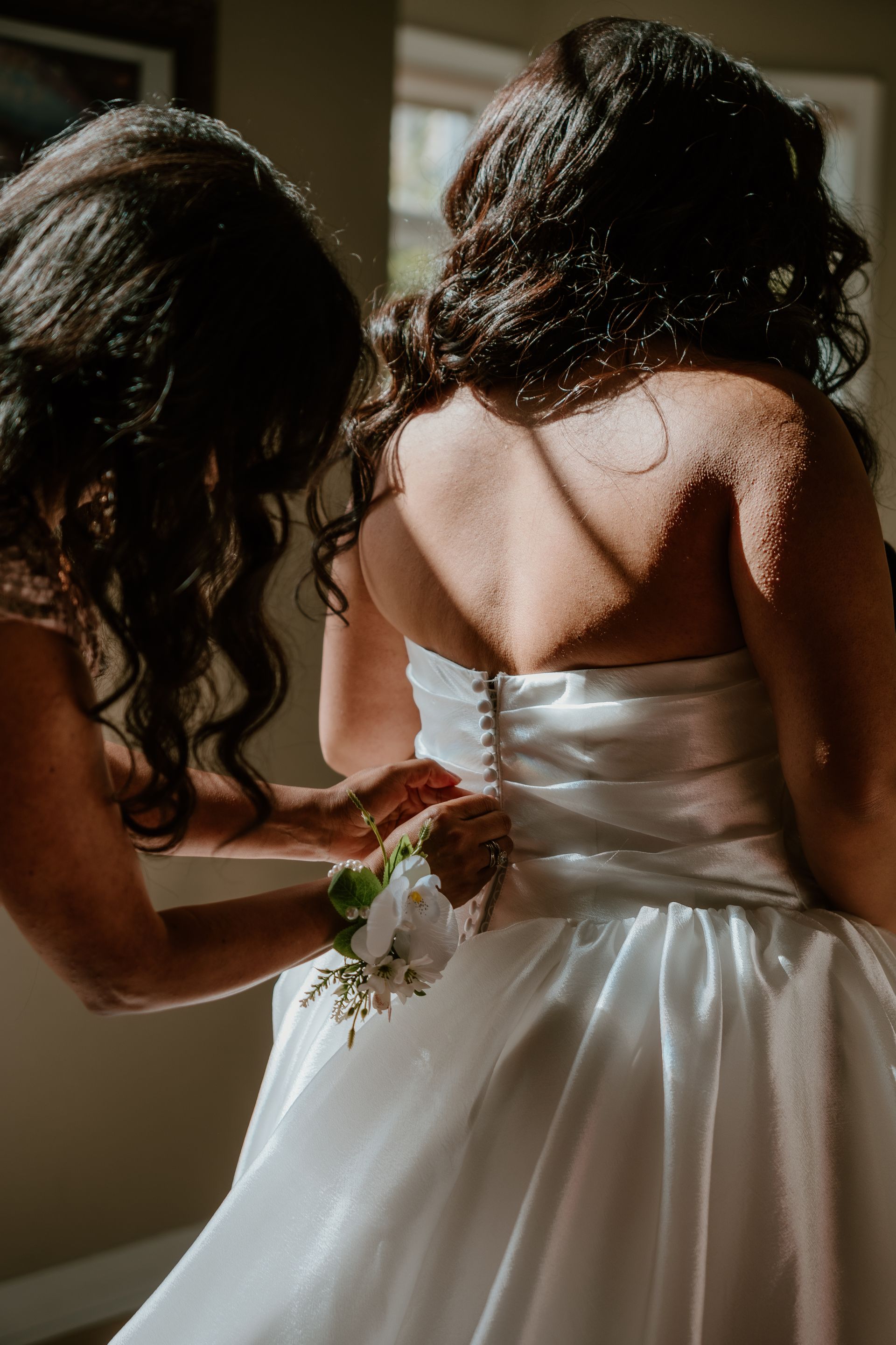 Bridal attendant fastening a white wedding dress on a woman in warm indoor light