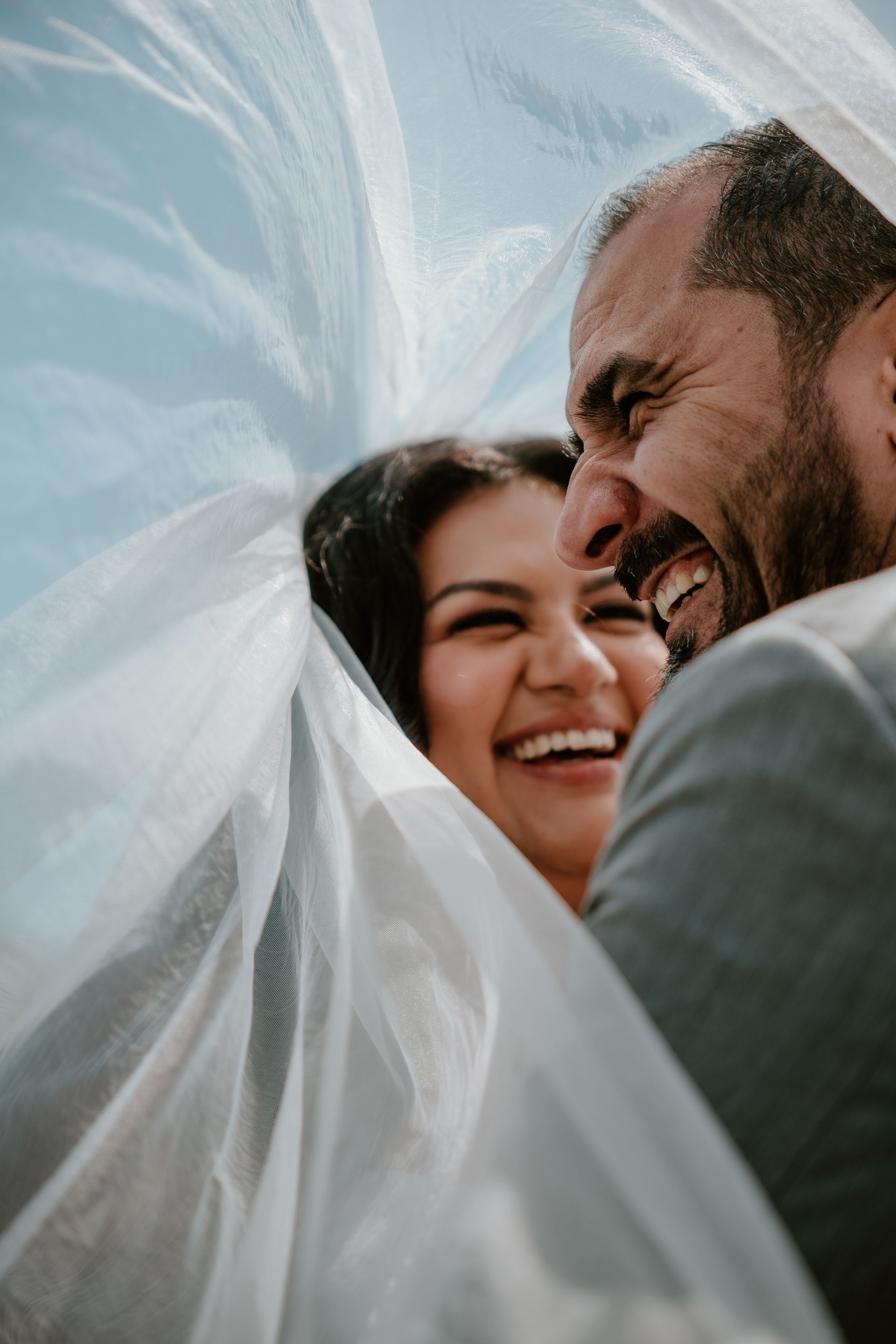 Smiling couple wrapped in a white veil, sharing a joyful close-up embrace