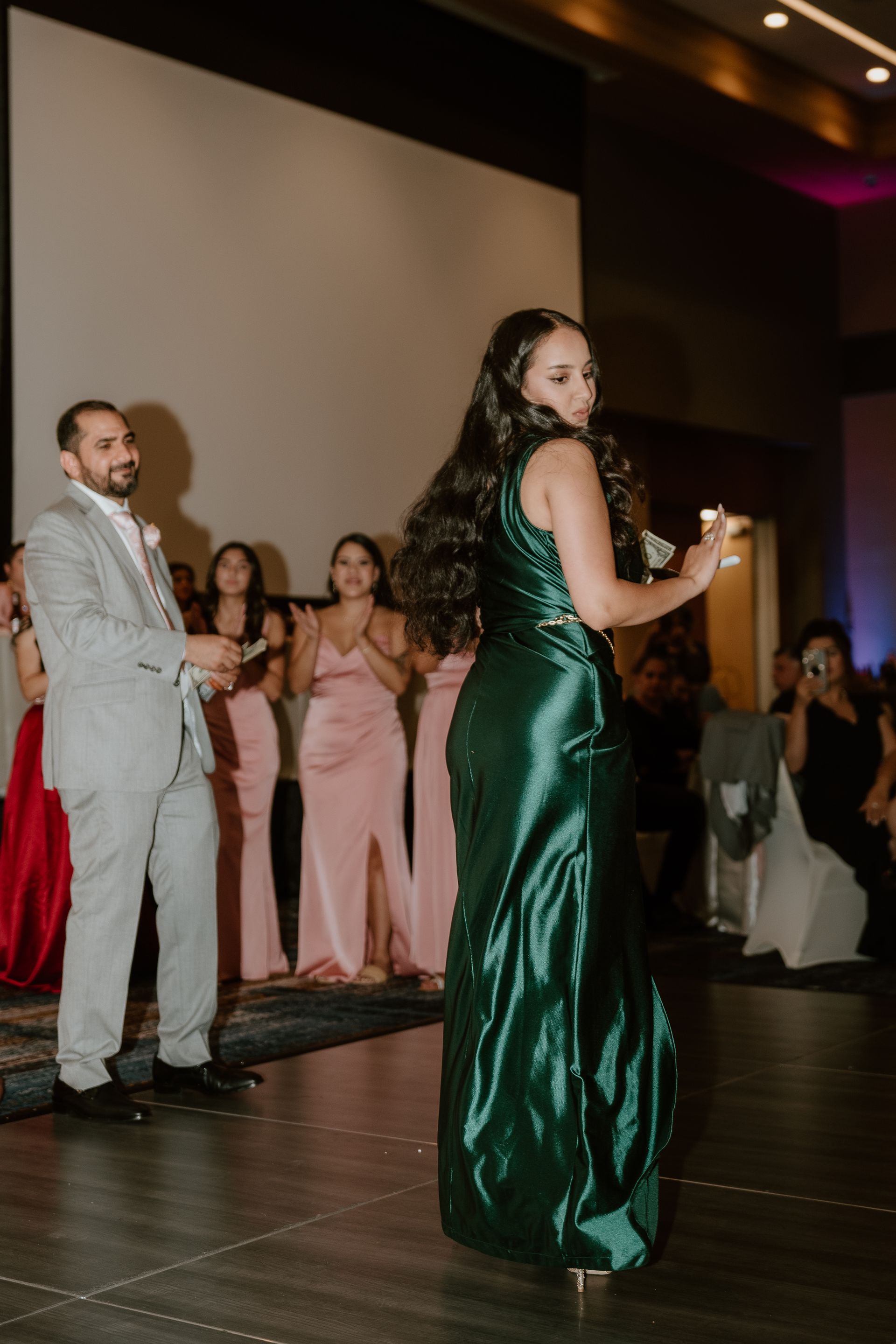 Woman in emerald green gown holding a microphone at a formal indoor event, with guests watching behind her.
