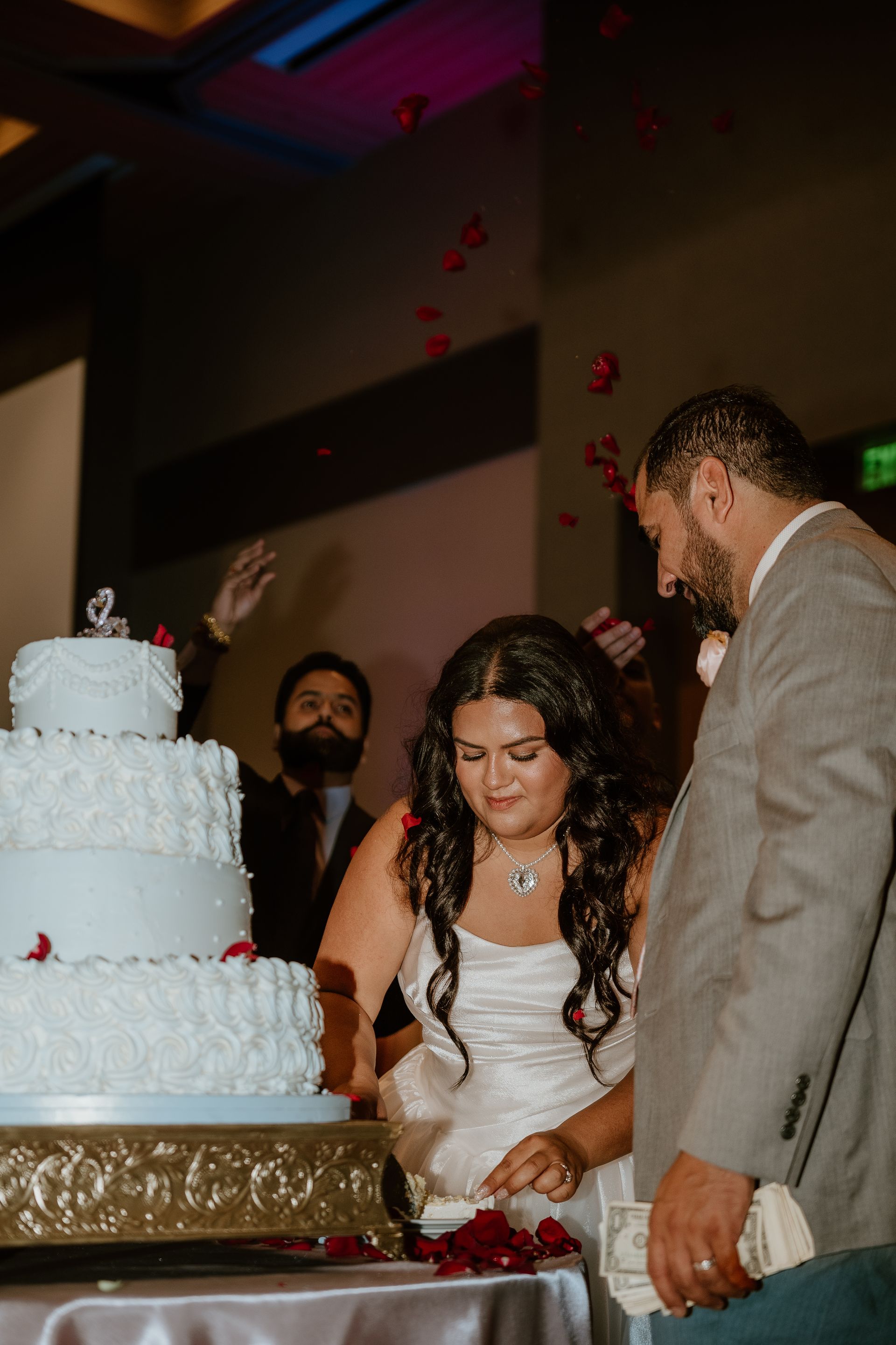 Couple cutting a white tiered wedding cake at a reception table, with a guest nearby and petals falling.