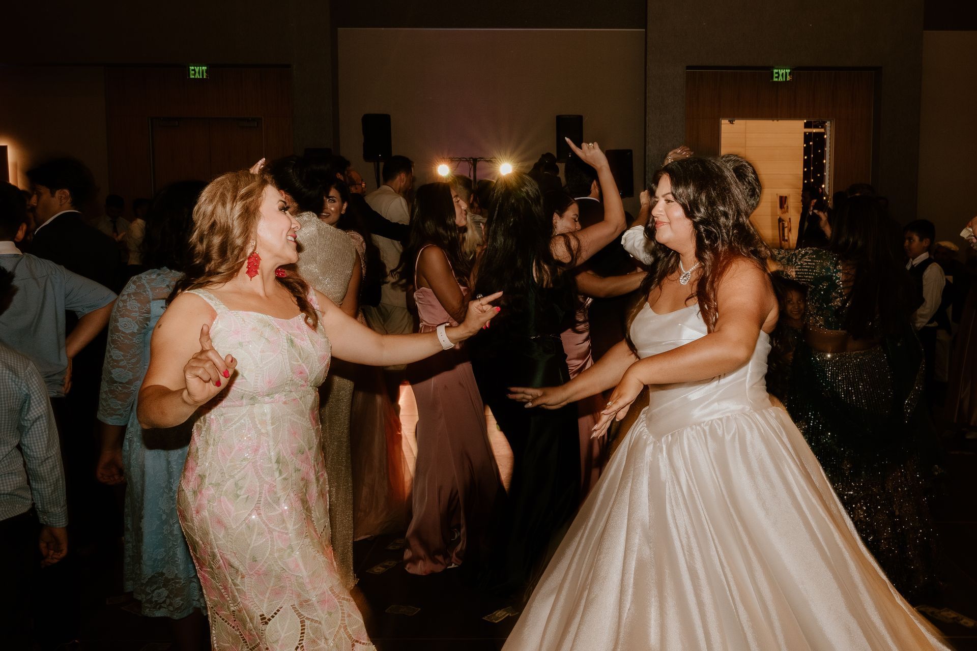 Two women in white gowns dancing at a formal reception, with guests and warm lights in the background