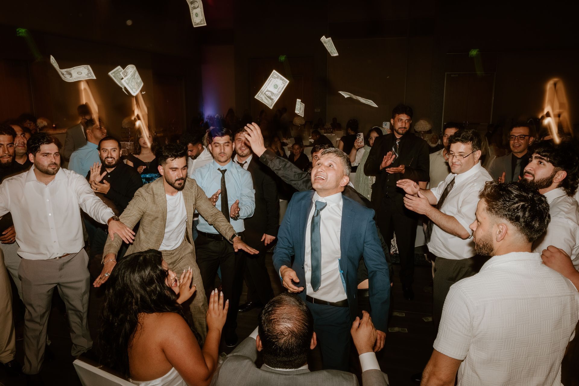 People dancing and cheering at a lively indoor party, with a man in a blue suit centered in the crowd.