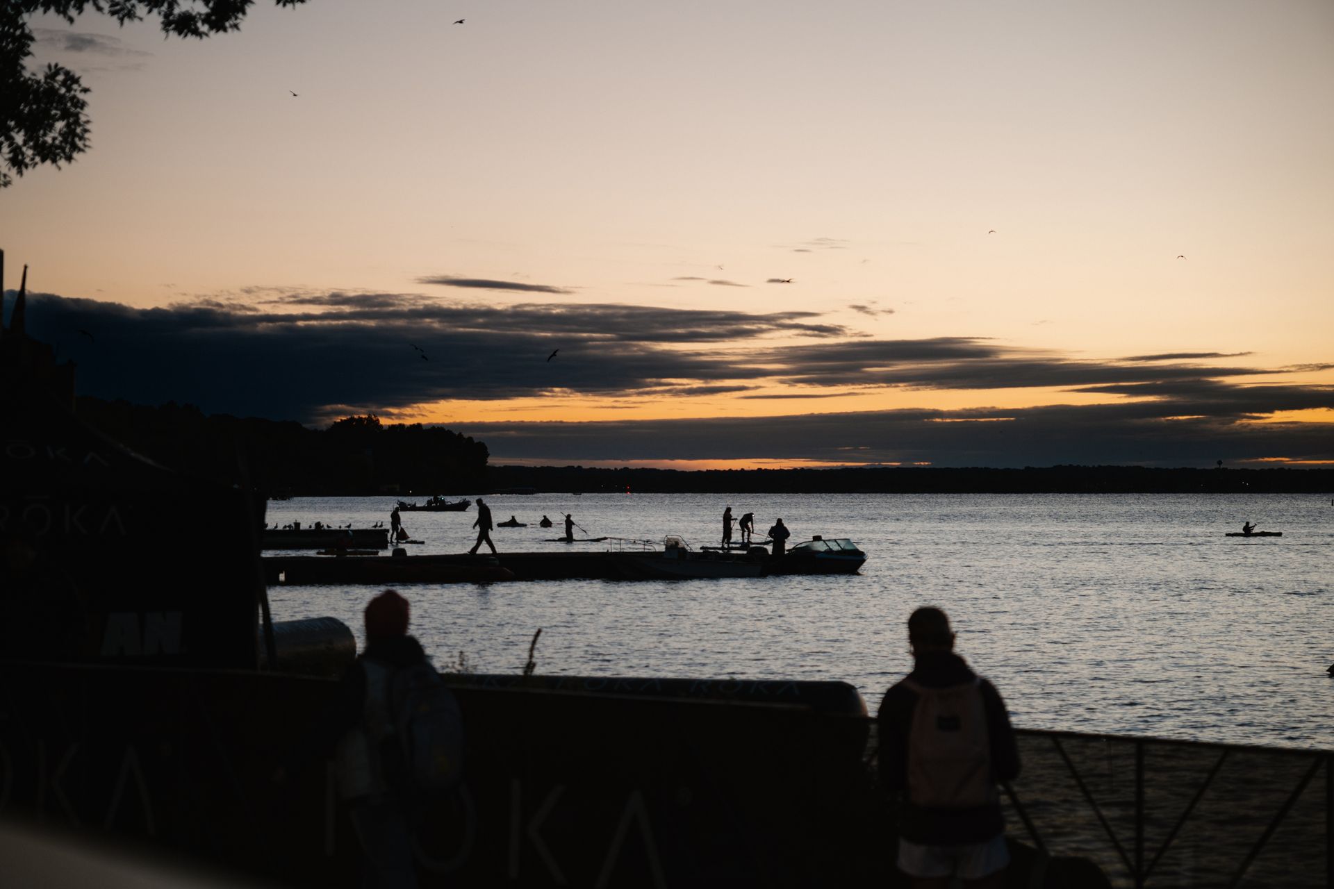Silhouetted people on a pier at sunset over calm water, with orange sky and dark clouds