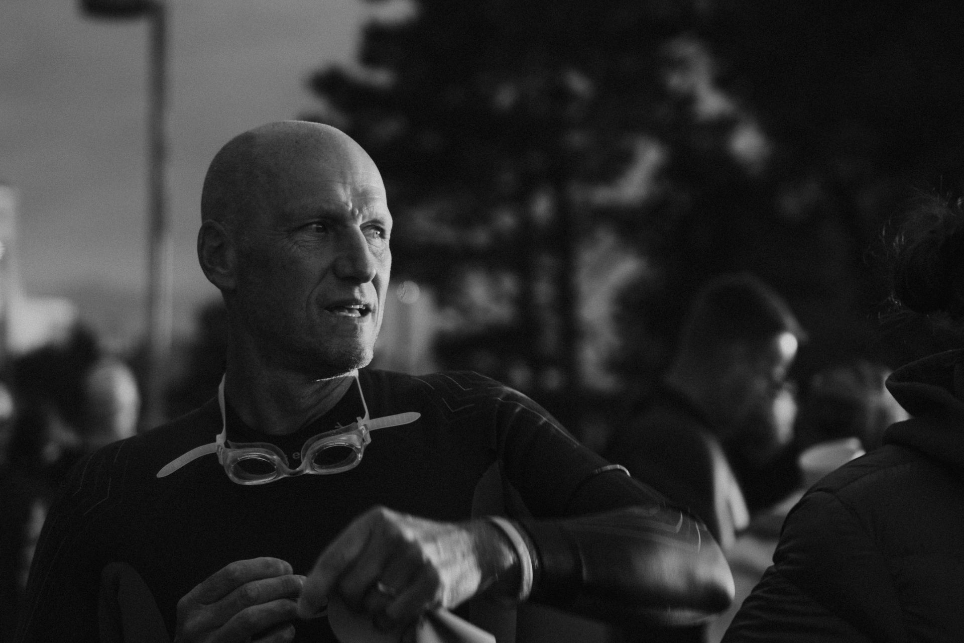 Black-and-white portrait of a man in a wetsuit, holding goggles at a crowded outdoor event
