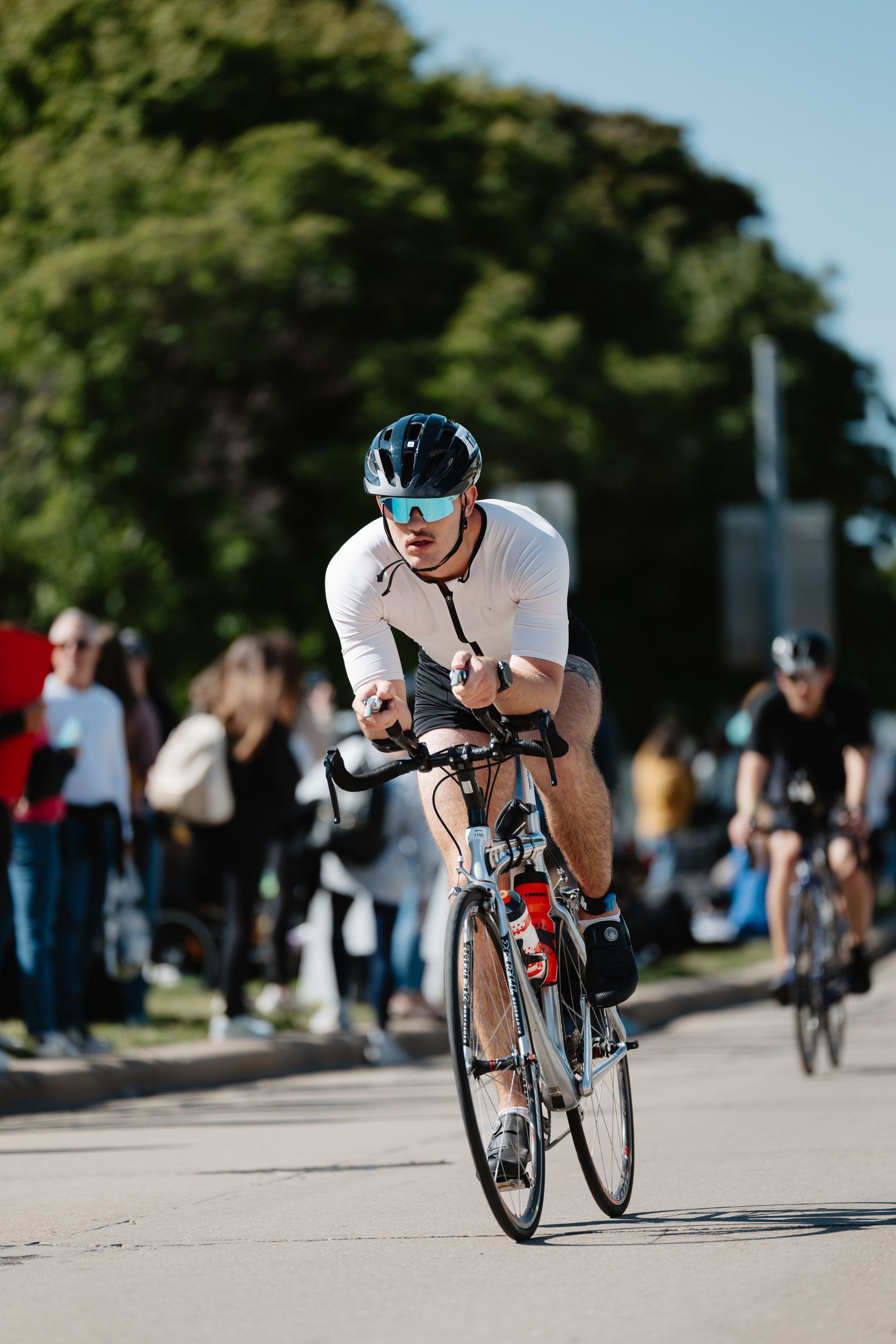 Cyclist in white jersey and blue helmet racing on a road with other riders and spectators behind