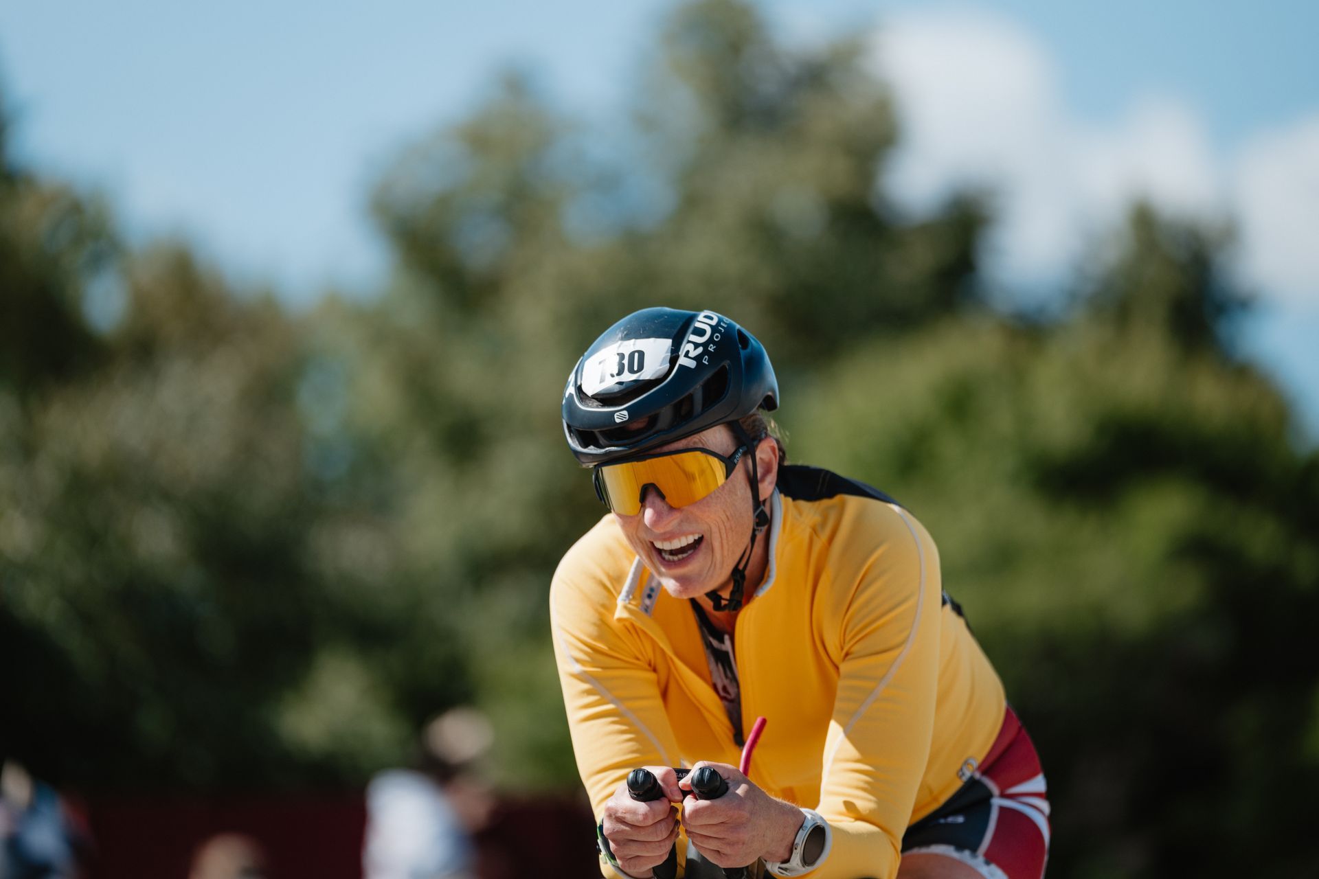 Cyclist in yellow jersey and helmet racing outdoors, leaning forward against a blurred tree background