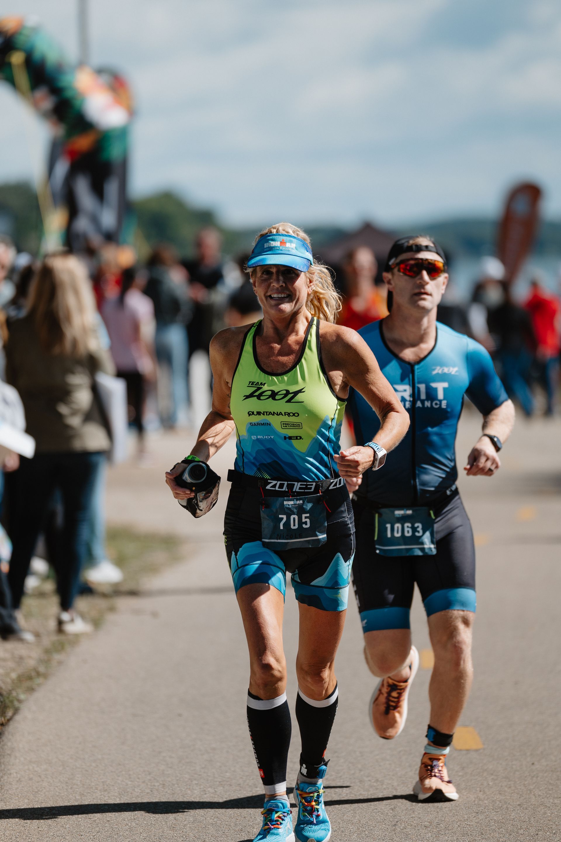 Two runners in blue and green racing on a road with spectators in the background.