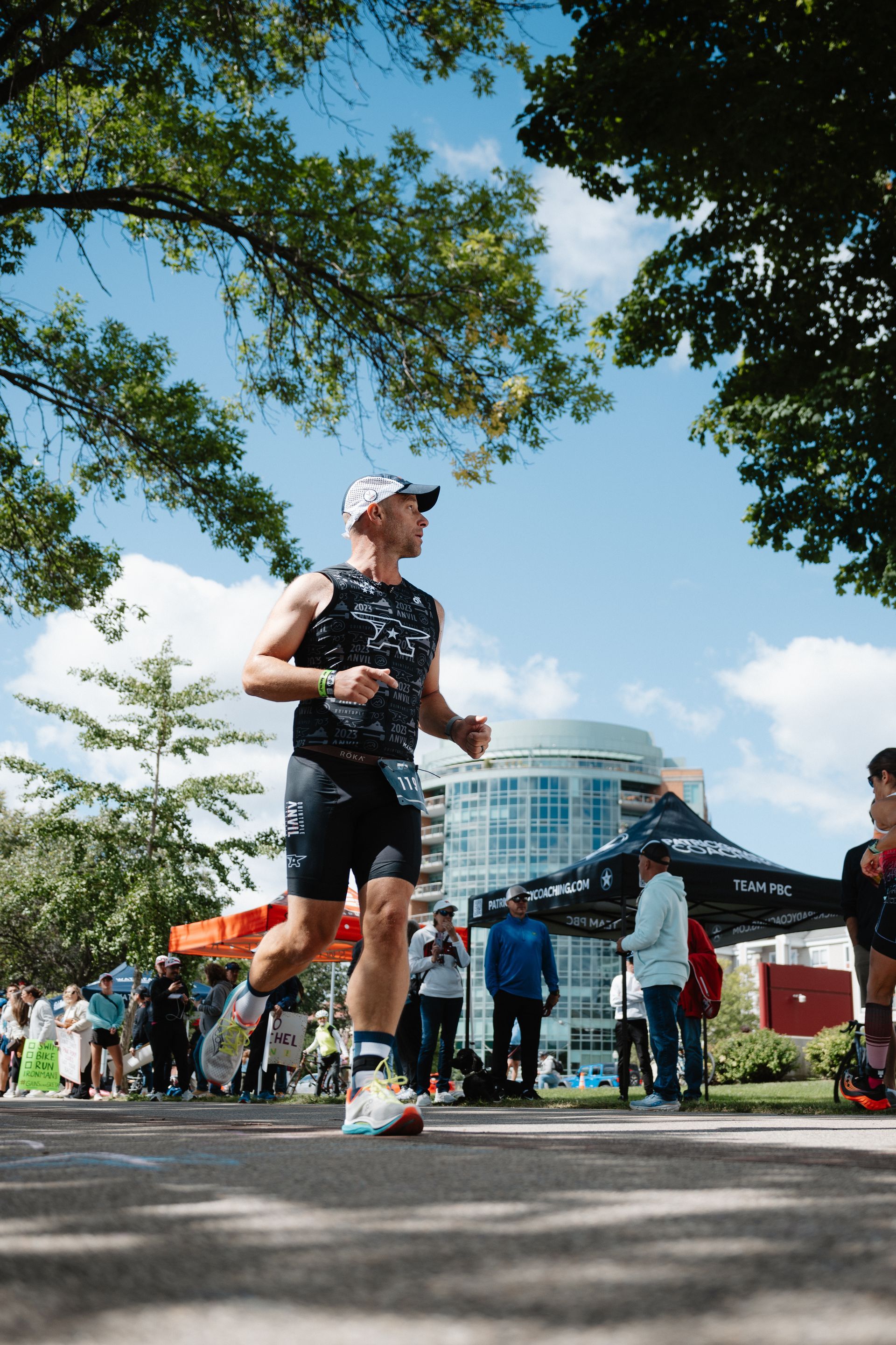Runner in black athletic gear jogging past tents at an outdoor race in a park under trees and blue sky