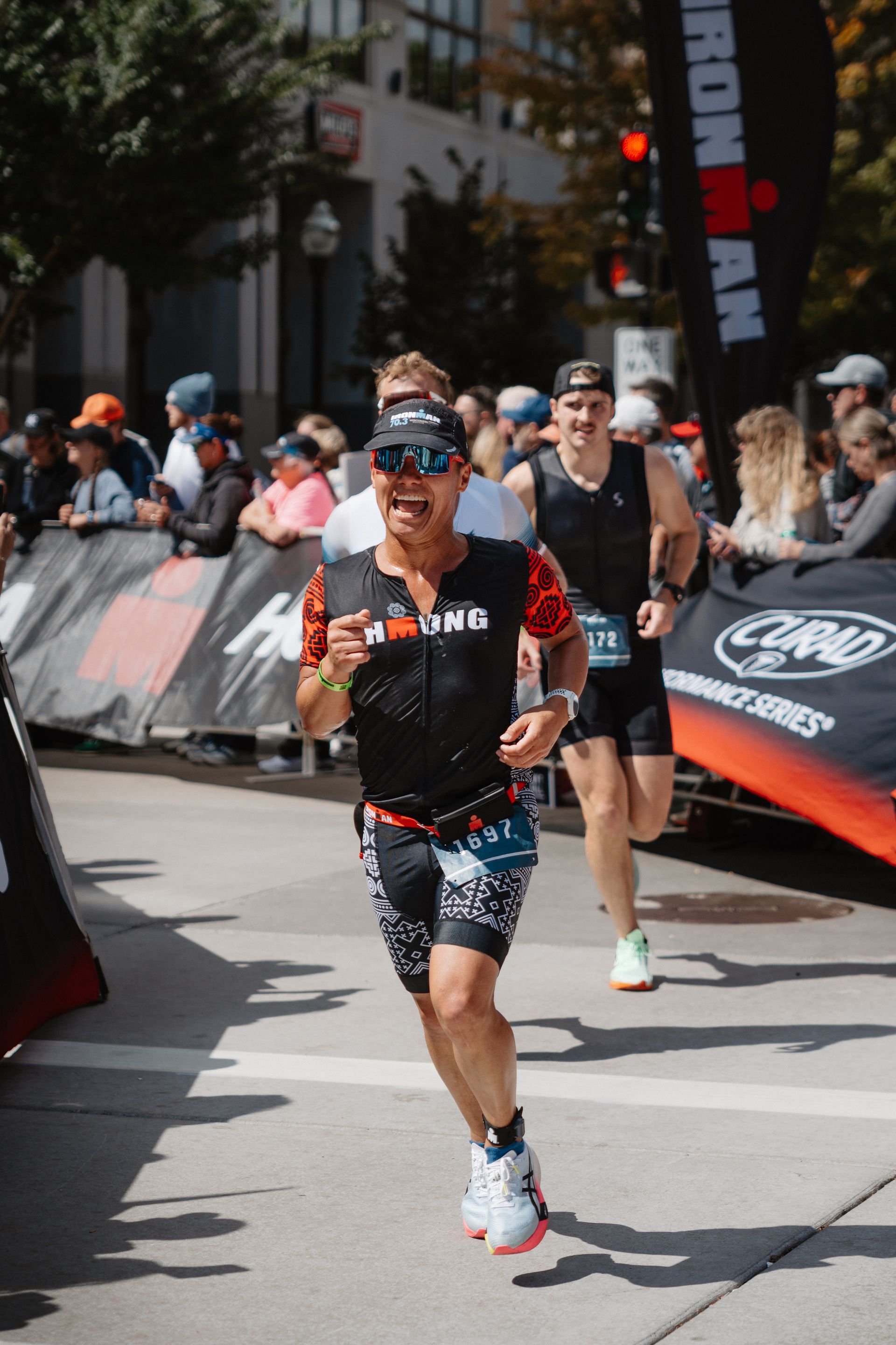 Runner in black-and-orange race gear crossing a finish area on a city street, with spectators behind barriers.